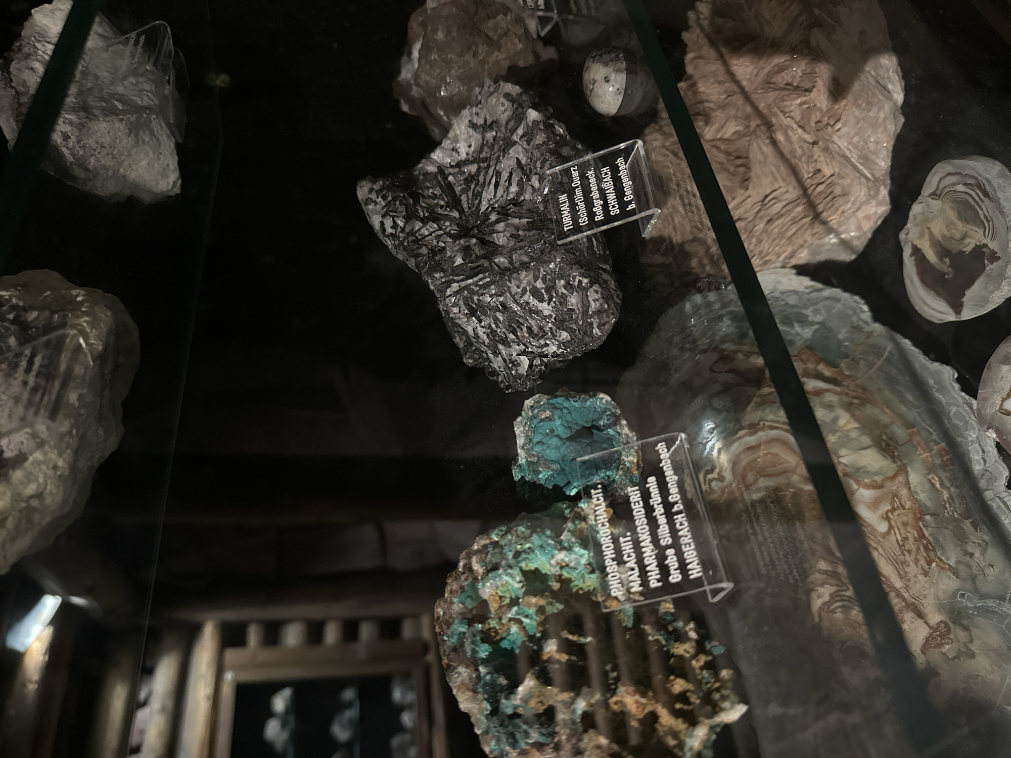 This image shows a collection of mineral specimens displayed in a museum or exhibition. The minerals are housed in glass cases, with some specimens resting on dark surfaces. The lighting is dim, creating a dramatic effect that highlights the textures and colors of the rocks.
Several distinct mineral samples are visible. One prominent specimen is a black and white crystalline rock with sharp, radiating black needles, labeled "TURMALIN (Schörl) im Quarz Roßgrabeneck. SCHWAIBACH b. Gengenbach". Another sample is a vibrant turquoise-blue mineral, identified as "PHOSPHOROCHALCIT. MALACHIT. PHARMAKOSIDERIT Grube Silberbrünnle HAIGERACH b. Gengenbach". There are also other unlabelled mineral formations with layered, banded, and crystalline structures in shades of gray, brown, and white, suggesting a diverse geological collection.
In the background, wooden structural elements suggestive of a mine shaft or an older exhibition design are dimly visible, hinting at a connection to mining or geological history. The overall impression is one of a carefully curated display of natural wonders, inviting viewers to appreciate the intricate beauty of mineral formations. The text labels provide specific geological information, including mineral names, compositions, and origins, suggesting the collection may be from the Schonach im Schwarzwald region of Germany.