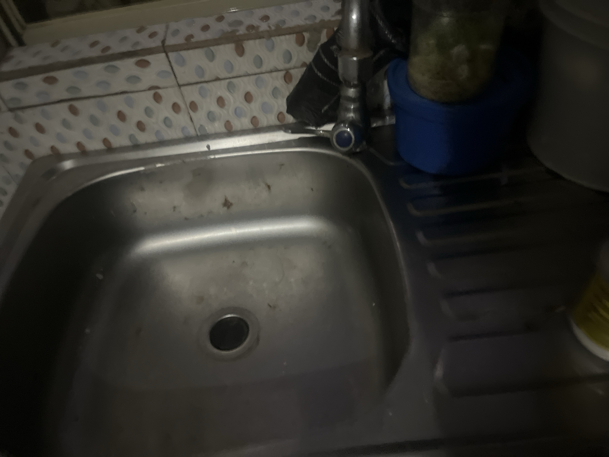 This is a close-up, slightly blurry, overhead shot of a stainless steel kitchen sink. The sink has a drain at the bottom center. To the left of the sink, there is a patterned tile backsplash with circular and oval shapes. Above the sink, a chrome faucet with a blue handle is visible. To the right of the faucet, a blue container sits on the counter, with a glass jar containing green contents placed on top of it. Further to the right, a portion of a gray object, possibly a bowl or container, is visible. The counter appears to be made of the same stainless steel material as the sink, featuring a ribbed drainage area to the right of the sink basin. A yellow bottle is partially visible on the far right edge of the frame. The lighting is dim, casting shadows and obscuring some details. The scene is likely a domestic kitchen. This scene is not specifically identifiable as being in Jalingo, Nigeria, based solely on the visual information provided.