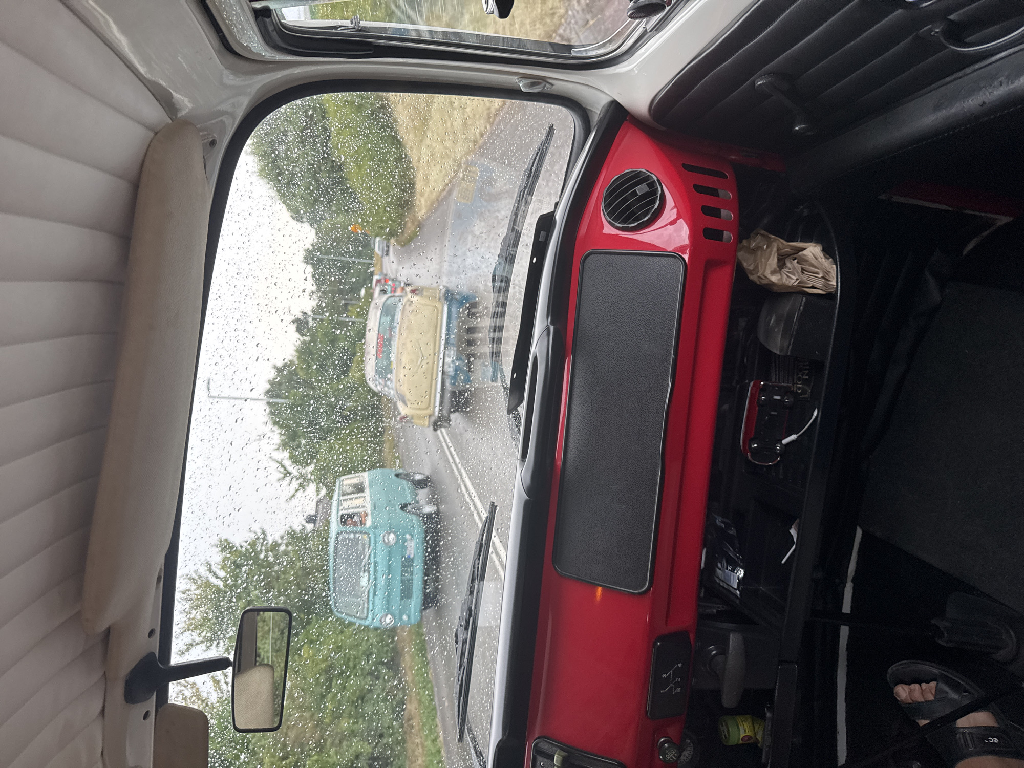 The image is taken from inside a vehicle, looking forward through the windshield. Raindrops are visible on the glass. The dashboard of the vehicle in the foreground is red with black accents and a black textured panel. A gear shift indicator with R, 1, 2, 3, and 4 is visible on the red dashboard. To the right of the red dashboard is a black vent. Below the dashboard is a black compartment with a brown paper bag and other items. A person's left foot wearing a black sandal is visible on the floor to the lower left.
Outside the vehicle, on a wet asphalt road, several cars are driving in a procession. The closest vehicle ahead is a yellow and blue vintage car. Further ahead is a turquoise vintage van. Trees and vegetation line the sides of the road. The sky appears overcast. The scene suggests a vintage car rally or parade.
The location is identified as Falkenberg, Sweden.