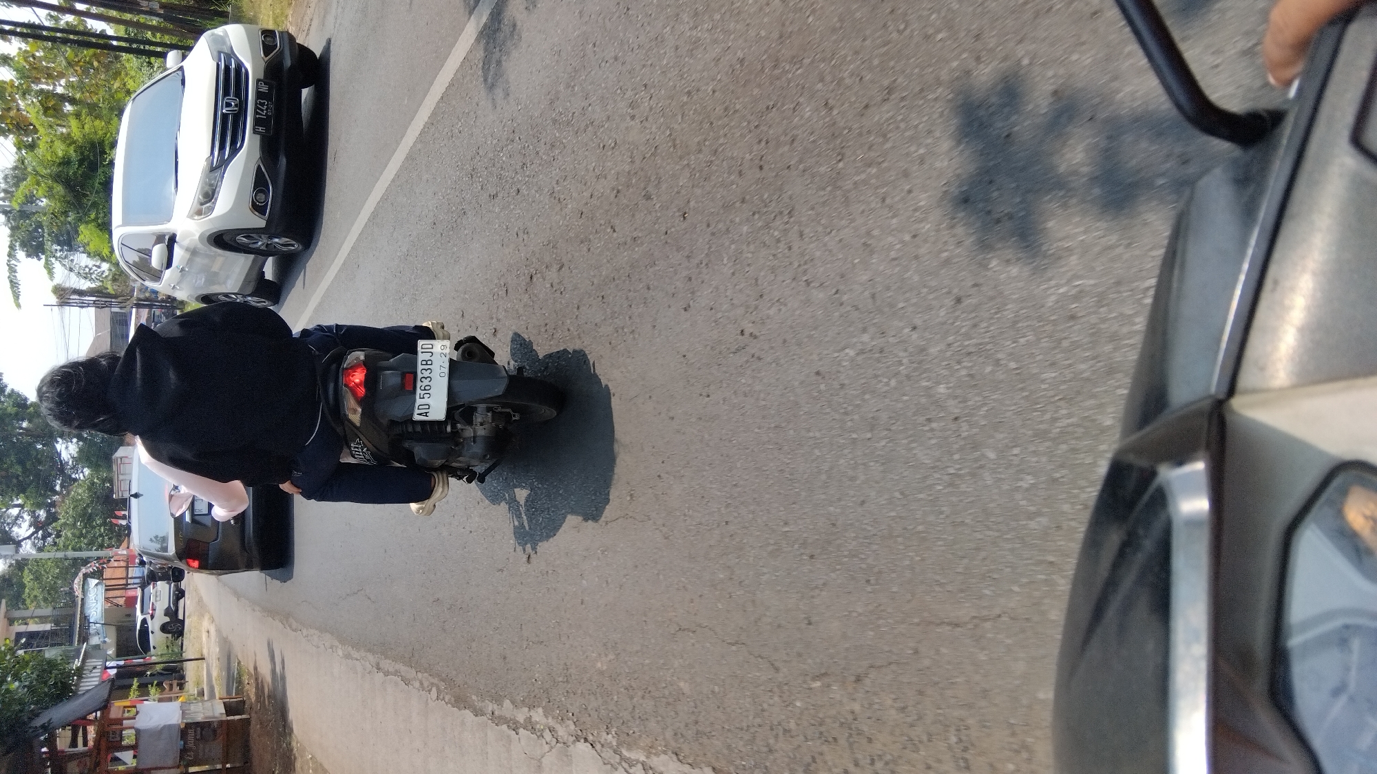 The image shows a street scene in Kalisalak, Indonesia, with a focus on a motorcycle carrying two people. The motorcycle has license plate "AD 5633 BJD". The front passenger is wearing a black hoodie and dark pants, while the rear passenger is wearing a pink shirt and dark pants. They are traveling on a paved road, with a white SUV to the right and a dark car ahead on the left. The environment appears to be urban or suburban, with buildings and trees visible in the background. The lighting suggests it is daytime.