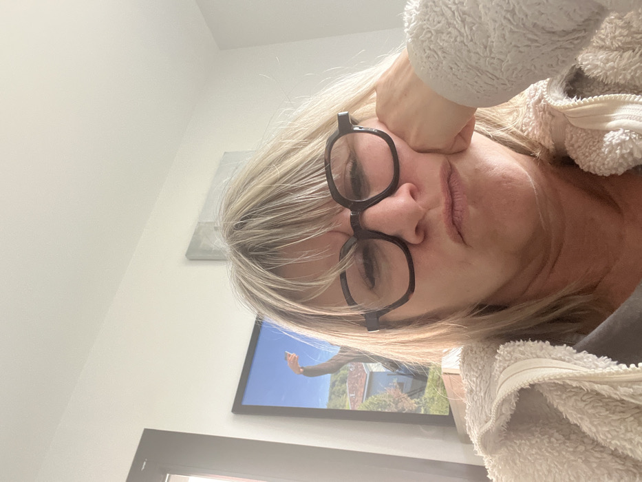 An indoor close-up photograph taken in Chambœuf, France, features an adult female centered in the frame. She wears dark-rimmed glasses and has blonde hair with bangs. Her expression is one of displeasure or boredom, indicated by furrowed brows, narrowed eyes, and pursed lips. Her left hand supports her chin. She is dressed in a light-colored, textured zippered jacket over a dark undershirt. The background is a plain white wall. To the subject's left, a framed artwork displays a landscape scene under a blue sky, with green foliage, a building featuring an orange-tiled roof, and a portion of what appears to be a human arm extending upwards. A second, smaller, rectangular, light-colored artwork hangs further right on the wall.