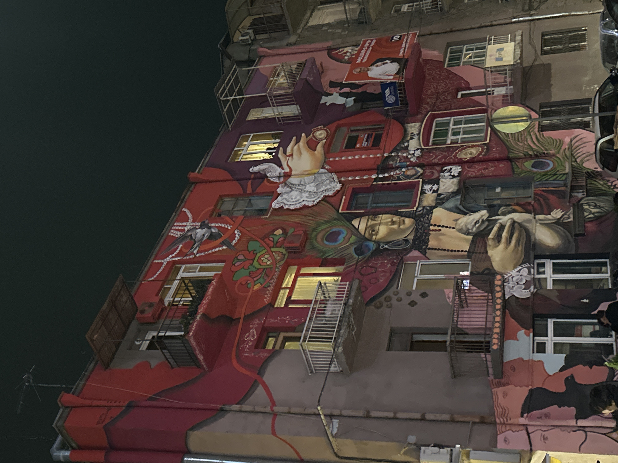 The image shows a tall building in Yerevan, Armenia, decorated with a large, colorful mural. The mural incorporates various elements, including portraits, hands, jewelry, peacock feathers, and abstract patterns, painted over the facade of the building. The building has windows and fire escapes, which are integrated into the artwork. The overall color scheme of the mural is predominantly red, pink, and brown. The background is dark, suggesting it is nighttime.