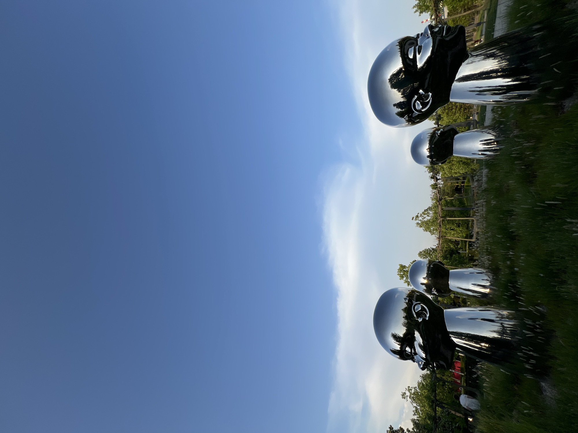 The image depicts a metallic sculpture in a grassy field, set against a backdrop of a clear blue sky with light clouds. The sculpture consists of human head forms, stacked vertically, appearing reflective. The scene is located in the city of Krasnodar, Russia. There is evidence of vegetation in the background, including trees. The picture is taken at an angle.