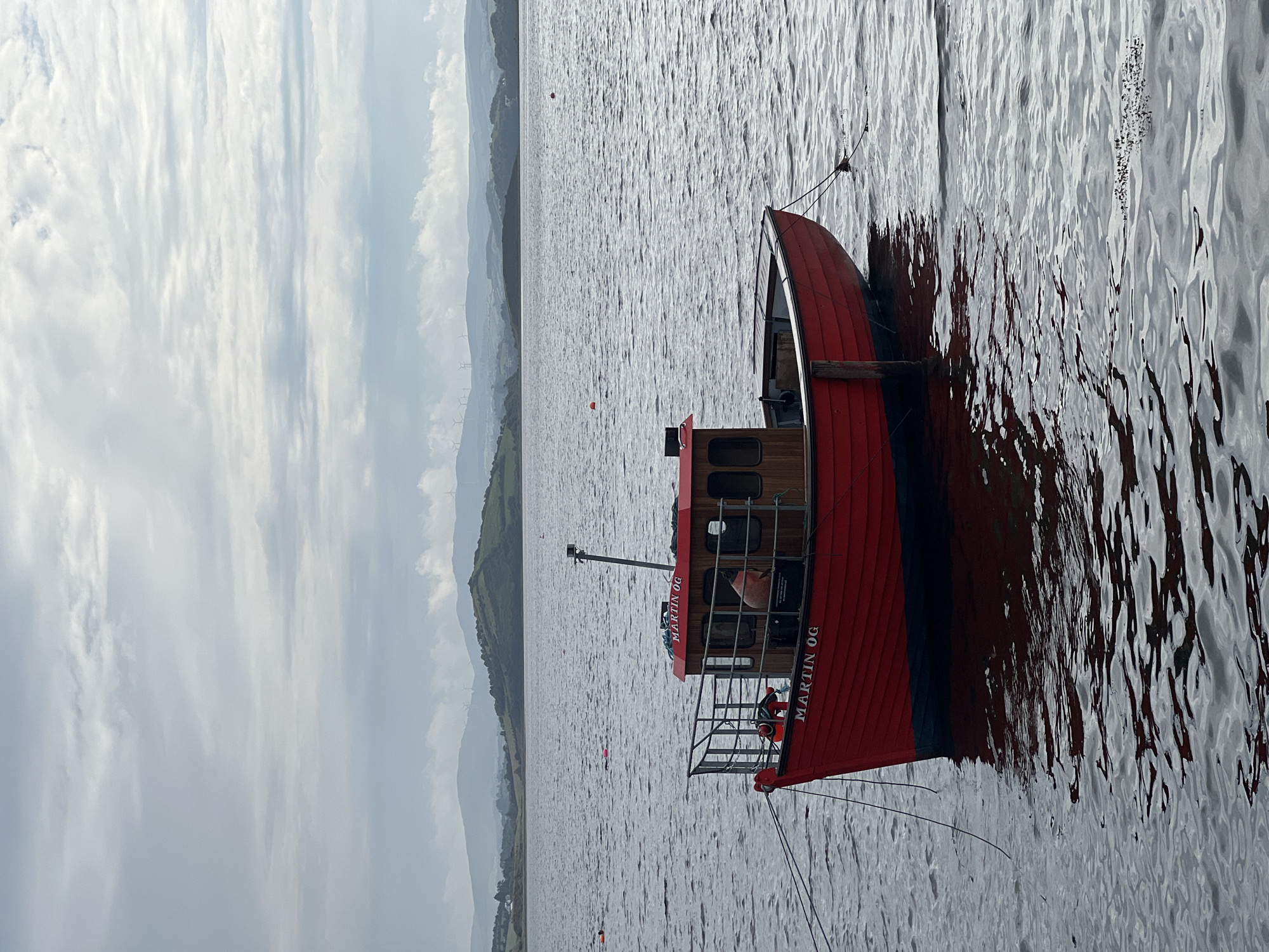 A red fishing boat named "MARTIN OG" is moored on a body of water. The boat has a wooden cabin and a metal railing. A person is partially visible inside the cabin. The water is choppy and reflects the overcast sky. In the background, a hilly landscape with scattered wind turbines can be seen. The scene appears to be set in a coastal area. No specific location within Terhillion, Ireland can be determined from the image.