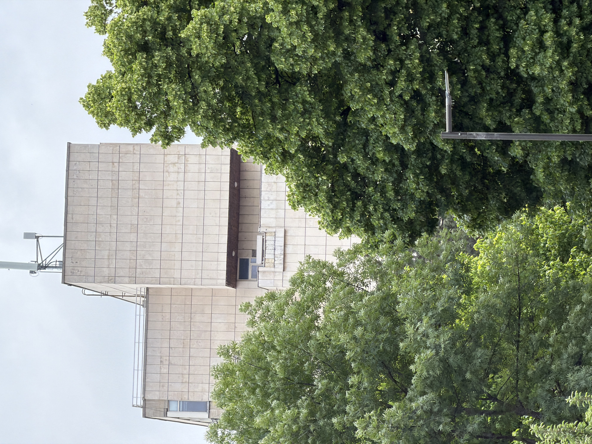 The image shows a building facade with a light beige or cream-colored tile pattern. A communication antenna is visible on the roof. Adjacent to the building, there is dense green foliage from trees, partially obscuring the view. A streetlight is visible between the trees. The angle of the building suggests that it's been rotated to the side. This appears to be the Bulgarian National Radio building located in Sofia, Bulgaria.