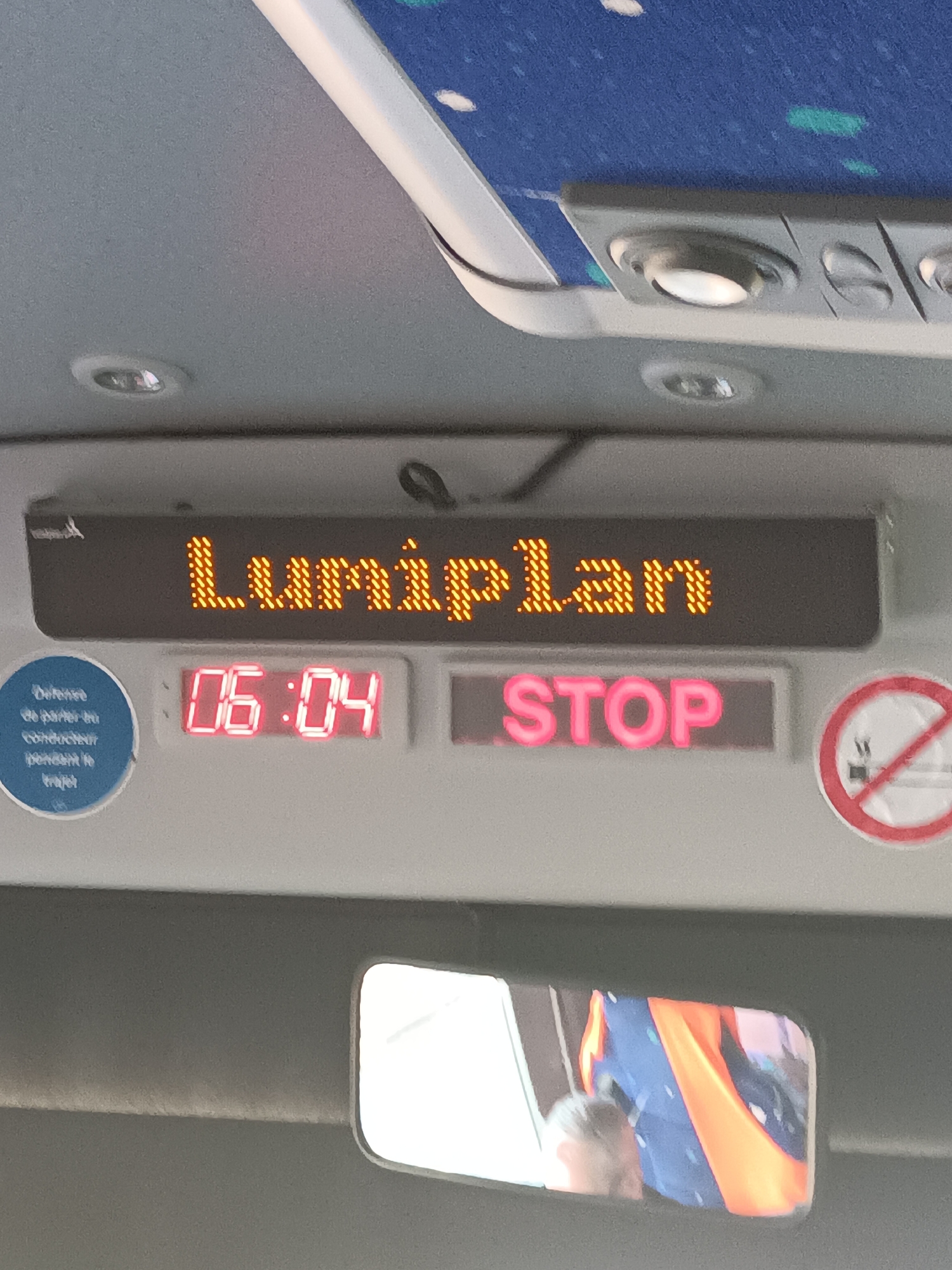 The image shows the interior of a public transportation vehicle, likely a bus or tram, in Canteleu, France. A digital display board shows the text "Lumiplan" in orange LED lights at the top. Below this, a red LED display shows the time "06:04". To the right of the time, a pink LED display shows the word "STOP". To the left of the time, there is a blue circular sticker with white text that appears to be an announcement. On the far right, there is a circular no-smoking sign with a red border. In the lower part of the image, a reflective surface, likely a mirror, shows a distorted reflection of people and colorful fabrics. The environment appears to be inside a moving vehicle, suggested by the interior lighting and the reflective surfaces.