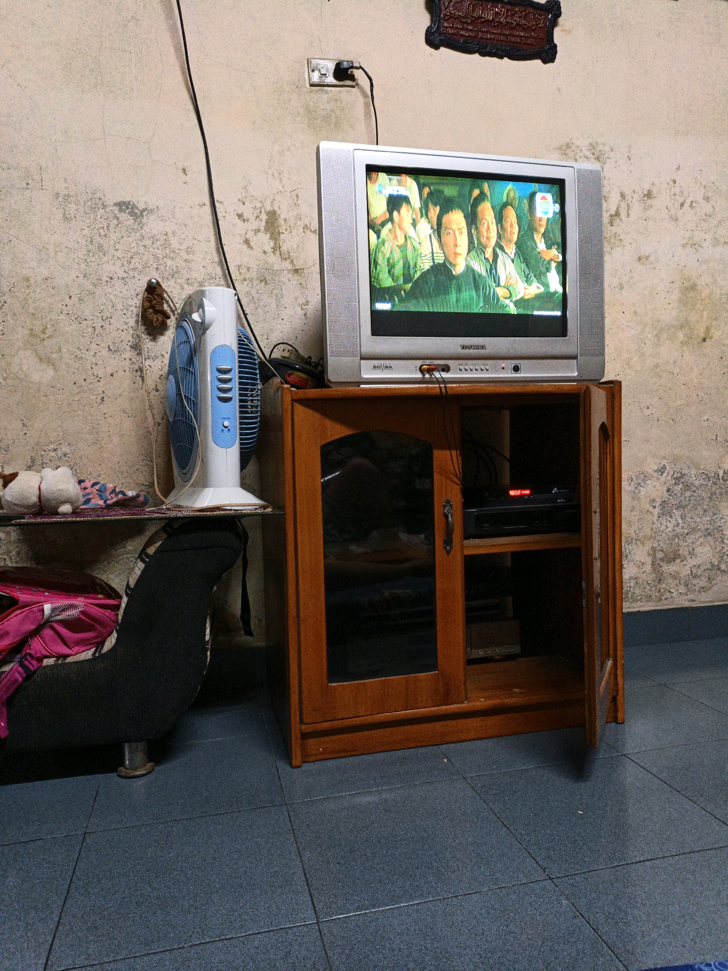 The image captures an indoor scene, likely a living area, with a vintage television set displaying a group of people. The television, a Toshiba, rests atop a wooden cabinet that has one door ajar, revealing some electronic equipment inside. To the left of the television, a blue and white standing fan is positioned near a shelf holding a small white stuffed animal and a patterned cloth. A black sofa, partially visible on the left, is covered with a pink and black bag. The walls are textured and appear aged, with visible cracks and discoloration. An electrical outlet with a plug is mounted on the wall above the fan. A decorative plaque with Arabic calligraphy hangs on the wall in the upper right corner. The floor is tiled in a dark blue hue. The overall impression is that of a simple, lived-in space. The presence of the fan, a television, and basic furniture suggests a domestic setting.