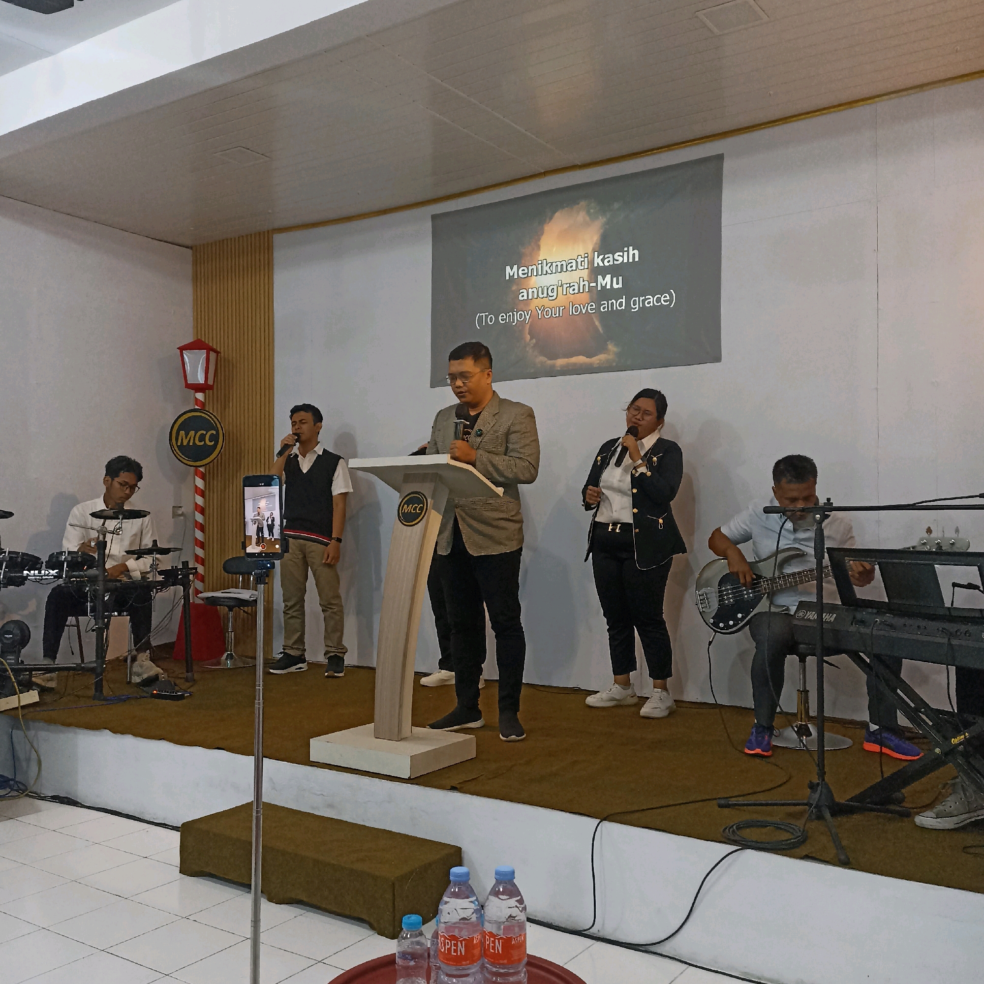 The image depicts a performance or religious gathering taking place on a raised stage within what appears to be a community hall or church in Magelang, Indonesia. At the center of the stage, a man wearing glasses and a gray blazer over a white shirt stands behind a wooden podium with the "MCC" logo. He is holding a microphone and speaking. To his left, a young man in a white shirt and black vest plays a drum set. To his right, a woman in a black blazer over a white shirt is singing into a microphone. Further to the right, a man in a light blue shirt plays a bass guitar while seated in front of a keyboard. A large screen behind the performers displays text in Indonesian and English that reads "Menikmati kasih anug'rah-Mu (To enjoy Your love and grace)". The stage is carpeted in brown, and the backdrop is a white wall with vertical wooden slats on the left side. A decorative red and white lamp post with a circular "MCC" emblem is positioned to the left of the stage. In the foreground, on a lower level, are two bottles of water and a smaller bottle of water on a red tray. The overall atmosphere suggests a religious service or a community event with live music.