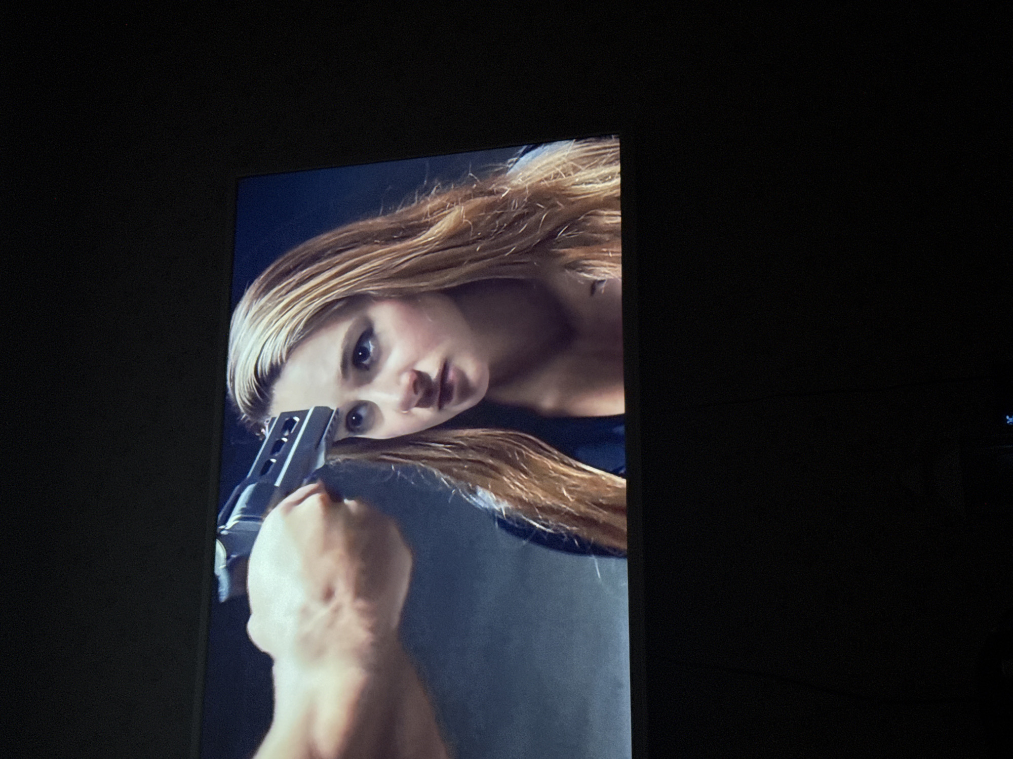 The image shows a television screen displaying a scene from a movie or TV show. In the scene, a woman with long, light brown hair is visible. A hand is holding a gun, aimed at the woman's head. The background is dark and indistinct. The image was taken indoors in a dark room. There is no information about the location in the city of Mytishchi, Russia.