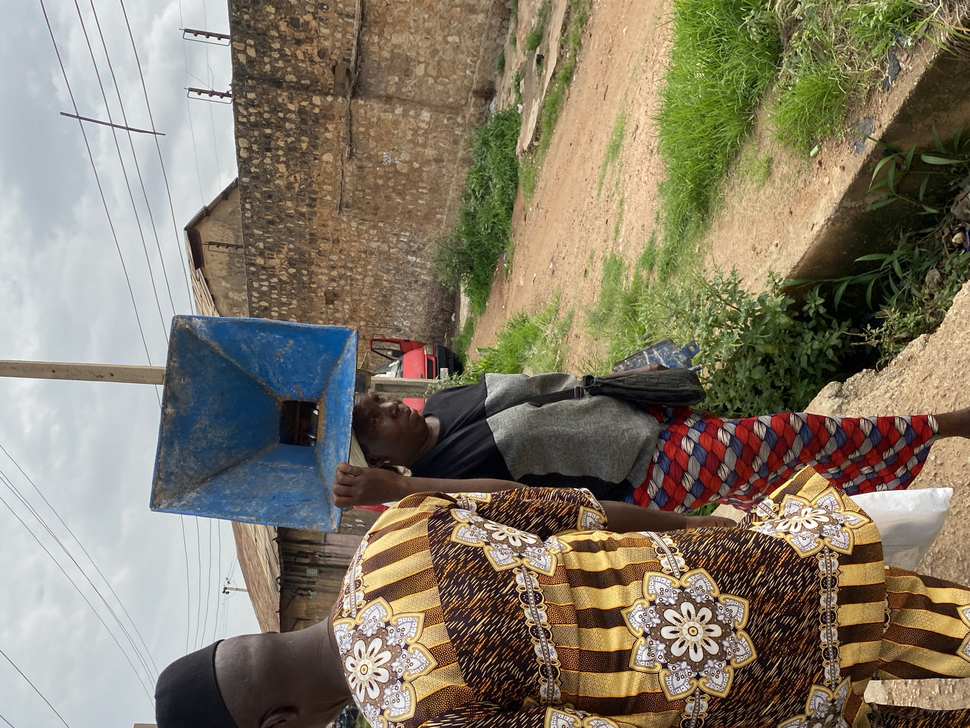 In Bukuru, Nigeria, two people are walking on a dirt path beside a concrete drainage ditch. The person in the foreground, seen from the back, is wearing a traditional dashiki with a pattern of yellow stripes and white floral designs. They have a black cap on their head and are carrying a white plastic bag in their left hand. To their right, a woman is walking and carrying a large, blue, funnel-shaped object on her head. She is wearing a dark shirt with a grey panel across the chest and a colorful skirt with a diamond pattern in red, white, and blue. She has a dark handbag slung over her shoulder. In the background, a stone wall runs along the right side of the path, with buildings made of corrugated metal and brick behind it. A red vehicle with its door open is visible on the left side of the image. The sky is overcast.