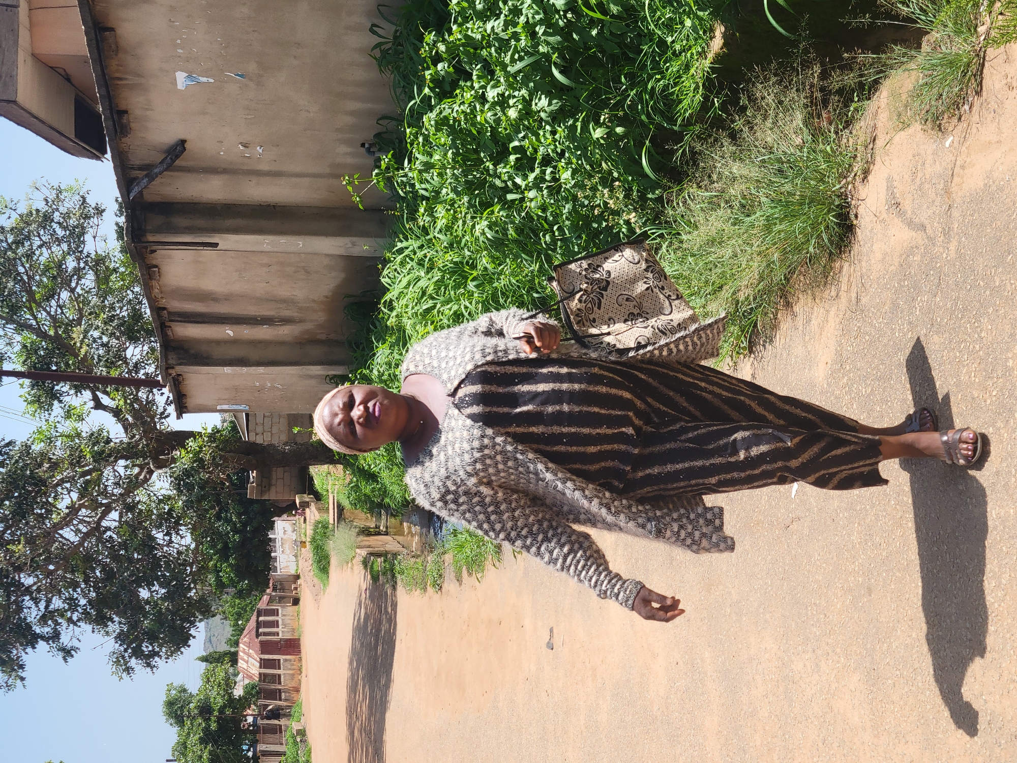 In Bukuru, Nigeria, a woman walks along a dirt road on a sunny day. She is wearing a light-colored, patterned cardigan over a dark, vertically striped dress and a light-colored headscarf. She carries a patterned handbag in her right hand. To her right, lush green vegetation grows along the edge of the road, bordering a drainage ditch. In the background, there is a light-colored wall with some peeling posters, a large tree, and a few simple buildings, suggesting a residential or semi-urban area. The road itself is dusty and uneven, with shadows cast by trees indicating the time of day.