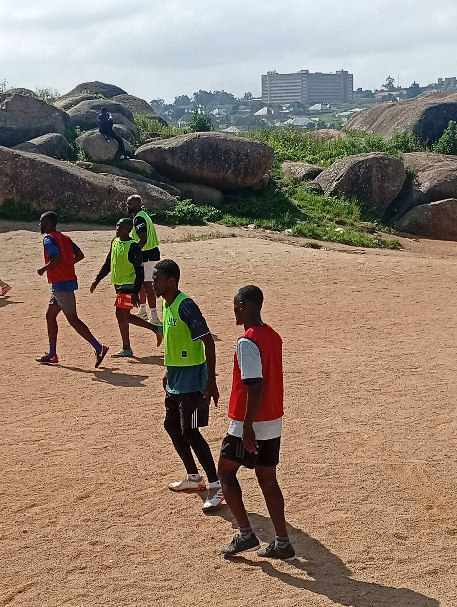The image depicts an outdoor scene in Jos, Nigeria, likely within the vicinity of the rock formations that characterize the city's landscape. In the foreground, several young men, appearing to be athletes, are engaged in some form of training or practice on a sandy or dirt surface. They are wearing athletic attire, including brightly colored bibs (red and lime green) over t-shirts and shorts. One individual in a red bib is running, while others appear to be standing or moving in a group. The background features large, weathered rock formations, typical of Jos. Beyond the rocks, a city skyline is visible, with buildings and some vegetation suggesting an urban or semi-urban environment. The sky is overcast with a hazy, bright appearance, indicating daylight. The overall impression is one of a sports activity taking place in a natural, rocky setting within the city of Jos.