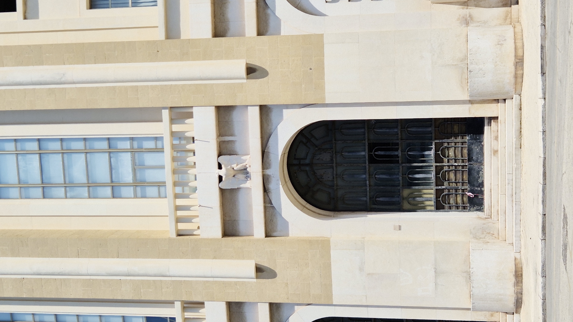 The image shows the entrance to a building, likely a church or public institution, with a large, arched, metal-grated door. The door has a dark, ornate design with multiple vertical elements that are curved at the top and bottom. Above the door, there is a white stone carving of an eagle with its wings spread. The building's facade is made of light-colored stone with vertical columns and large, rectangular windows with pale blue glass. A balustrade runs across the upper part of the facade. The entrance is accessed by a set of stone steps. The steps and the pavement in front of the building appear worn. A small section of asphalt road is visible in the foreground. A pink ribbon is tied to the door. The overall impression is of a grand, possibly historical, building. This building is the Chiesa di San Francesco di Assisi in Taranto, Italy.