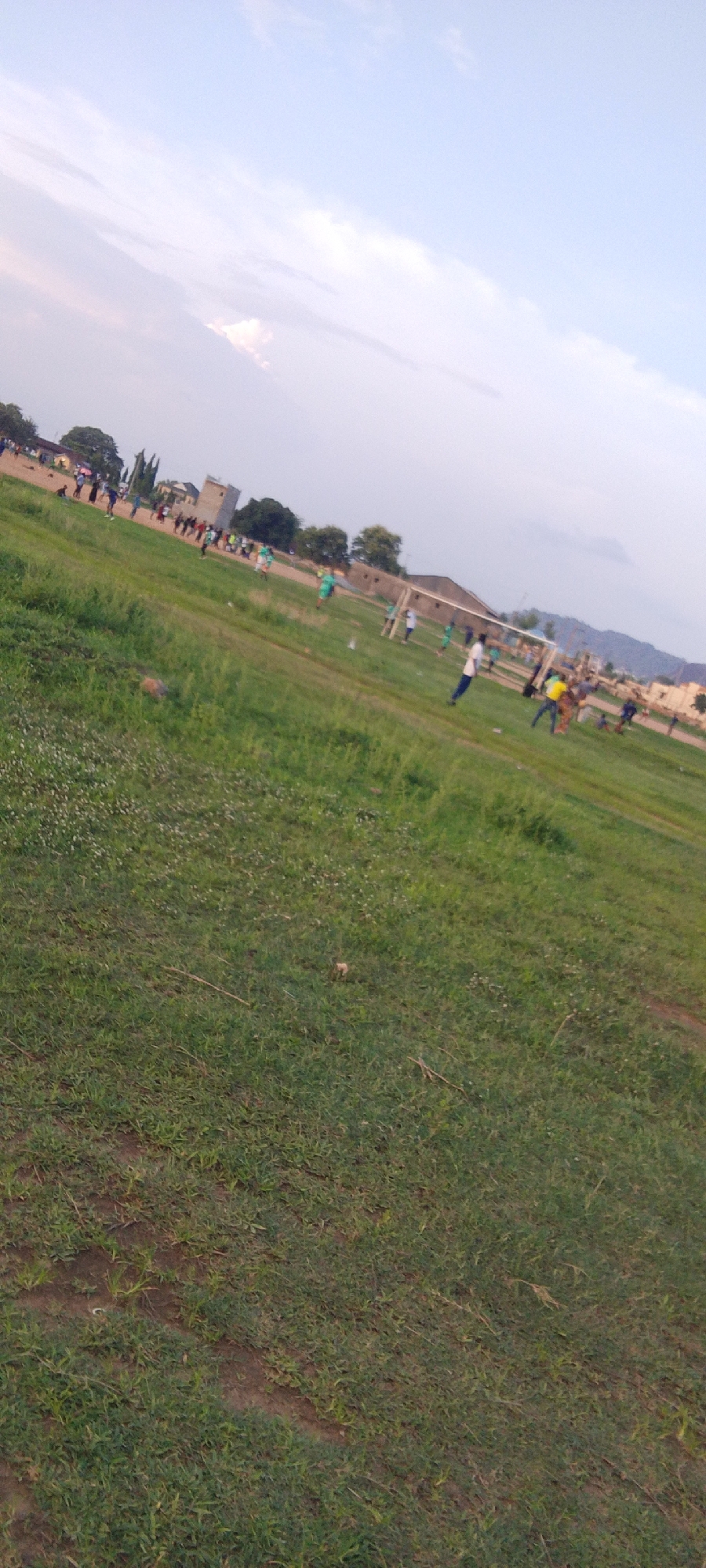 The image depicts a soccer game in progress on a grassy field, likely in a rural or semi-rural setting in or near Ibo, Nigeria. The field is uneven with some patches of bare earth visible in the foreground. A soccer goal is positioned in the mid-ground with players surrounding it, some wearing bright colored shirts. Other individuals are standing on the sidelines. In the background are buildings, trees, and a low mountain range. The sky is partly cloudy.