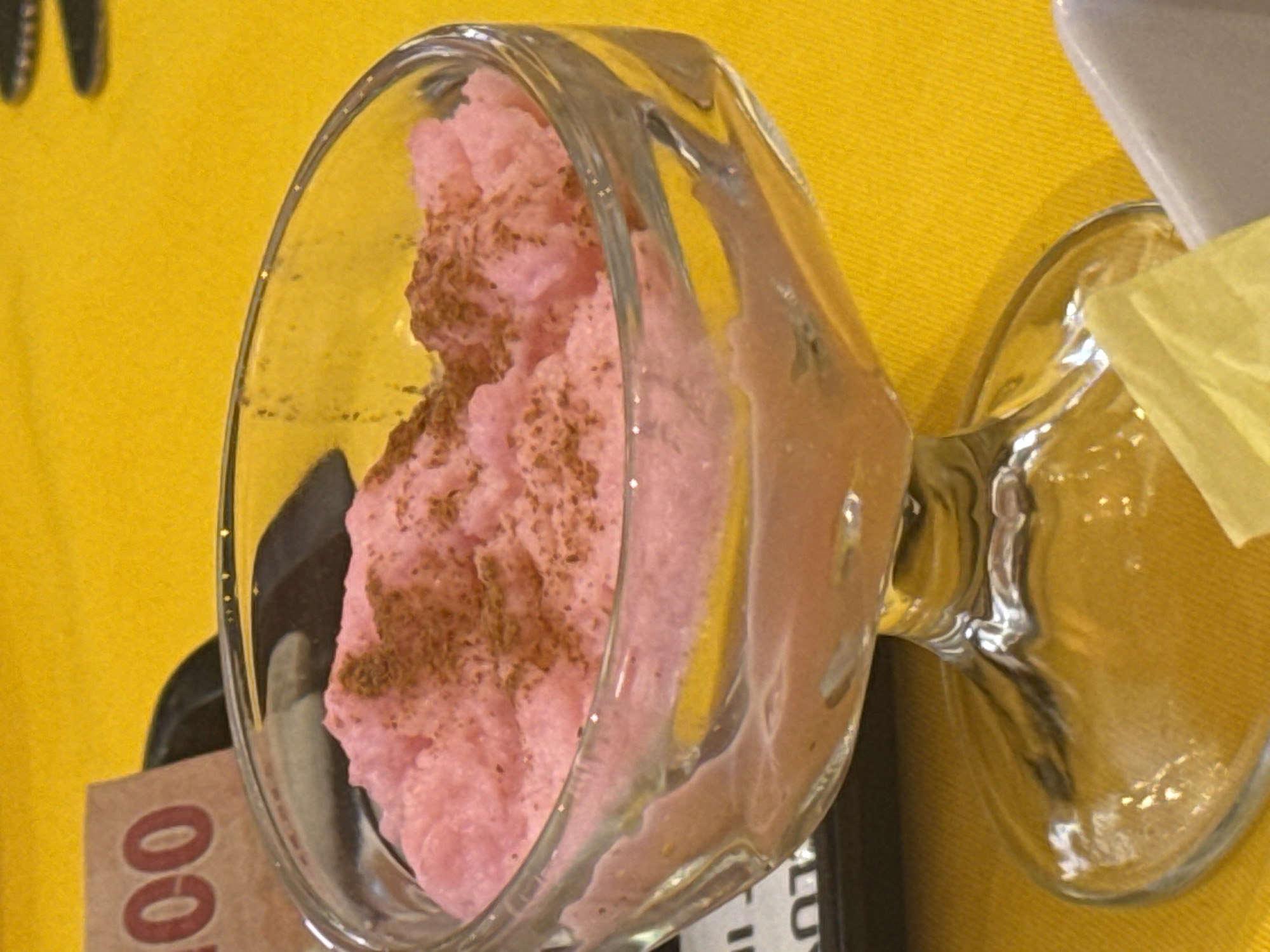A glass of pink dessert, possibly ice cream or mousse, dusted with brown powder, is on a yellow surface, possibly a tablecloth, in an indoor setting. The glass is partially transparent, and its contents are visible. Cutlery is vaguely visible in the background on the left. Other objects and surfaces are also present but not fully identifiable due to the shallow depth of field and composition. The scene does not provide any information to connect it to the city of San Cristóbal, Mexico.