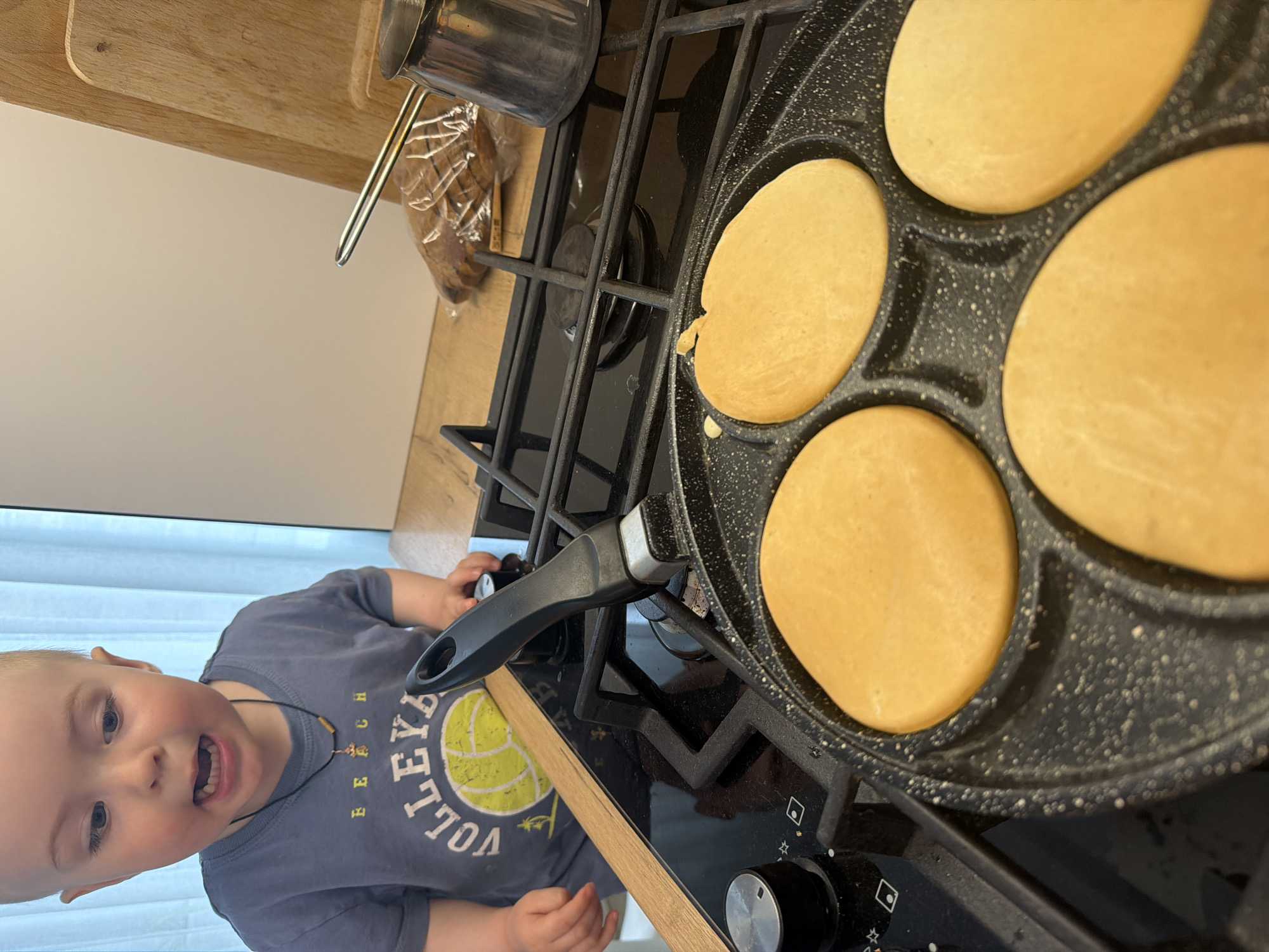 A toddler stands by a stovetop on which a multi-sectioned frying pan holds four pancakes. A small saucepan sits on the stovetop next to the pan, with a loaf of bread visible in the background. The location is undetermined, but it could be in the city of Votkinsk, Russia.
