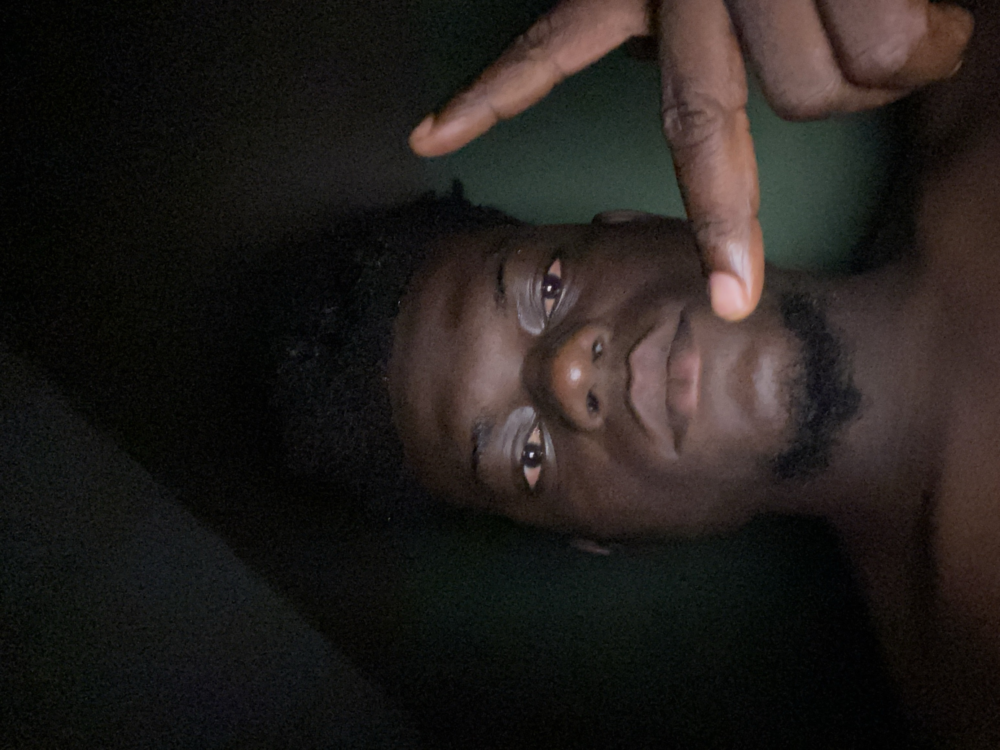 The image shows a dark-skinned individual, likely male, in a dimly lit environment. The person's face is the primary focus, with their eyes open and a serious expression. A hand is positioned above their face, with one finger pointing downwards. The background is dark and indistinct, making it difficult to discern any specific details about the environment. No information on the location, Unknown, Unknown, can be determined from the image.