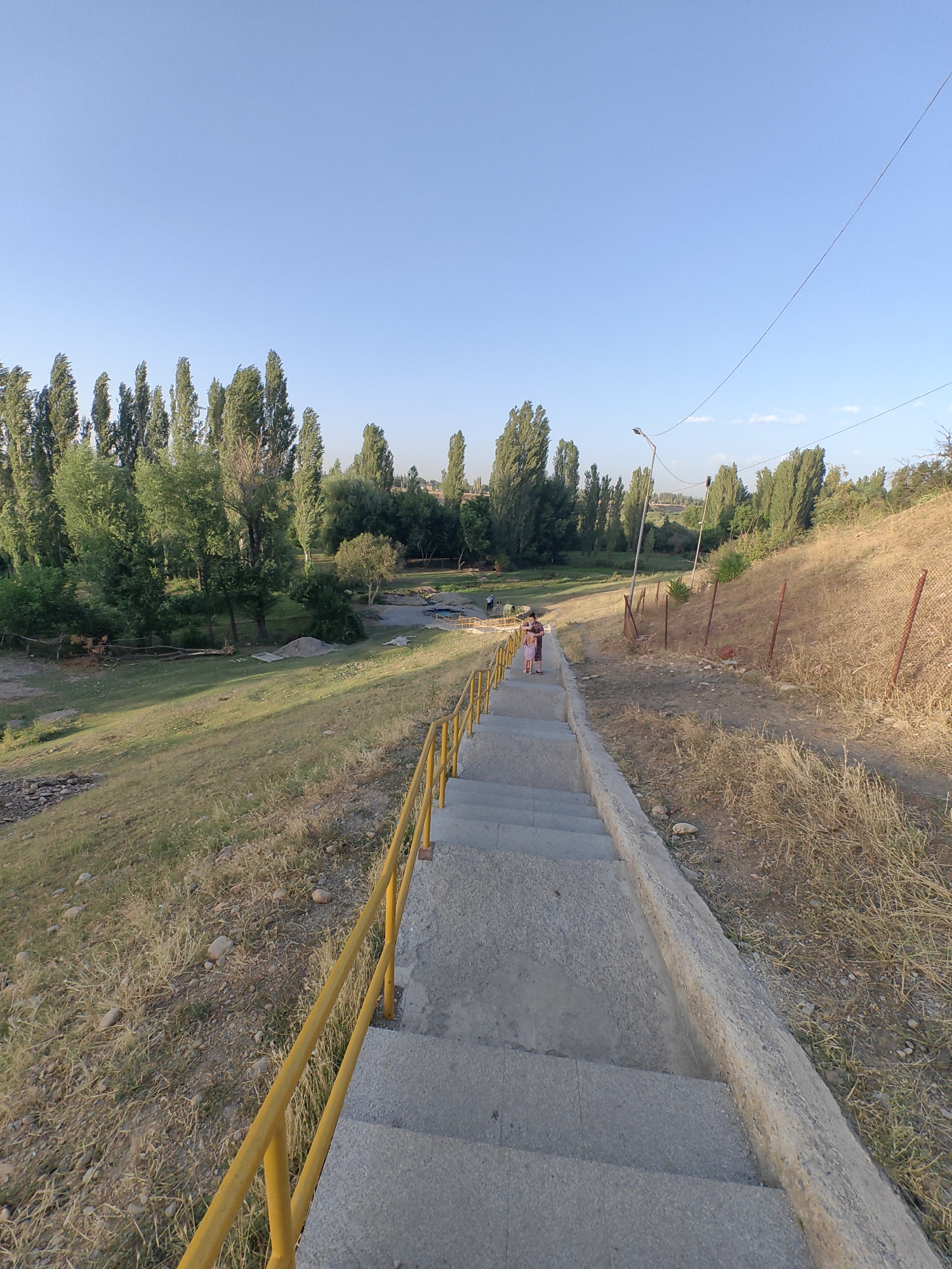 The image depicts a downward view of a concrete staircase with a bright yellow handrail on the left side, leading into a park-like area. The staircase is set against a sloping, grassy embankment covered with dry grass and scattered rocks. In the background, a cluster of tall, slender trees lines a body of water, and beyond that, more trees and a distant, hilly landscape are visible under a clear blue sky. Two individuals, possibly a child and an adult, are walking down the stairs. A lamppost stands on the right side of the path, and a wire is suspended diagonally across the upper right portion of the frame. The overall environment suggests a public outdoor space, likely within or near the city of Көксаяқ ауылдық округі, Kazakhstan.