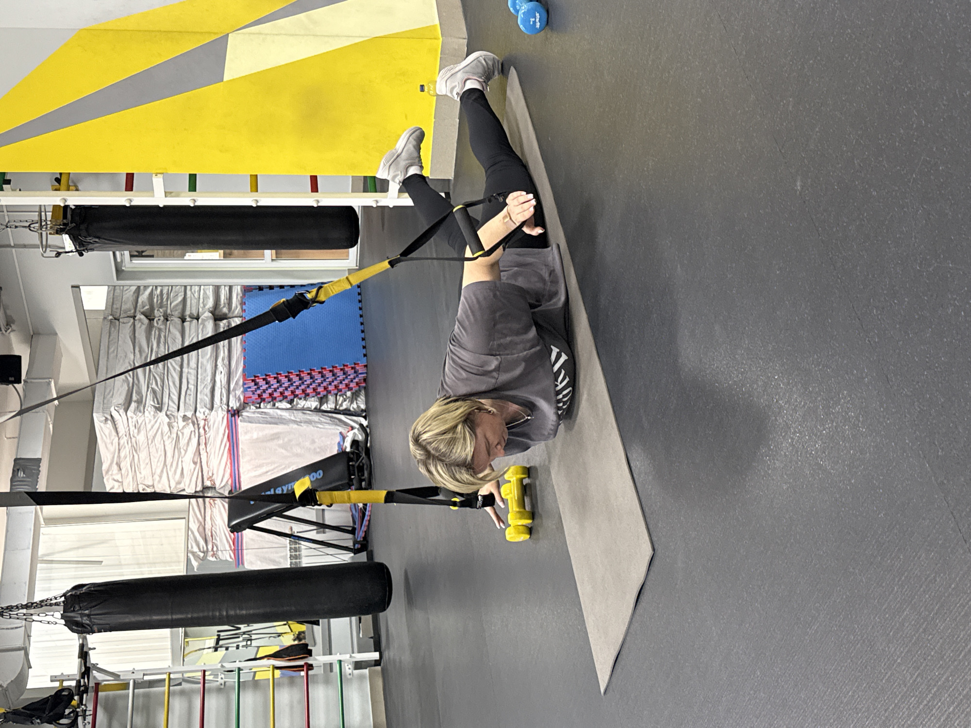 The image shows a woman exercising on a mat in a gym-like setting. She is using suspension straps attached to a frame overhead. There are punching bags and other gym equipment visible in the background. There are blue dumbbells next to the mat. The location appears to be a gym in an indoor setting, perhaps in the city of Chaikovsky, Russia, as indicated by the prompt. There are also yellow dumbbells near the woman's shoulder.