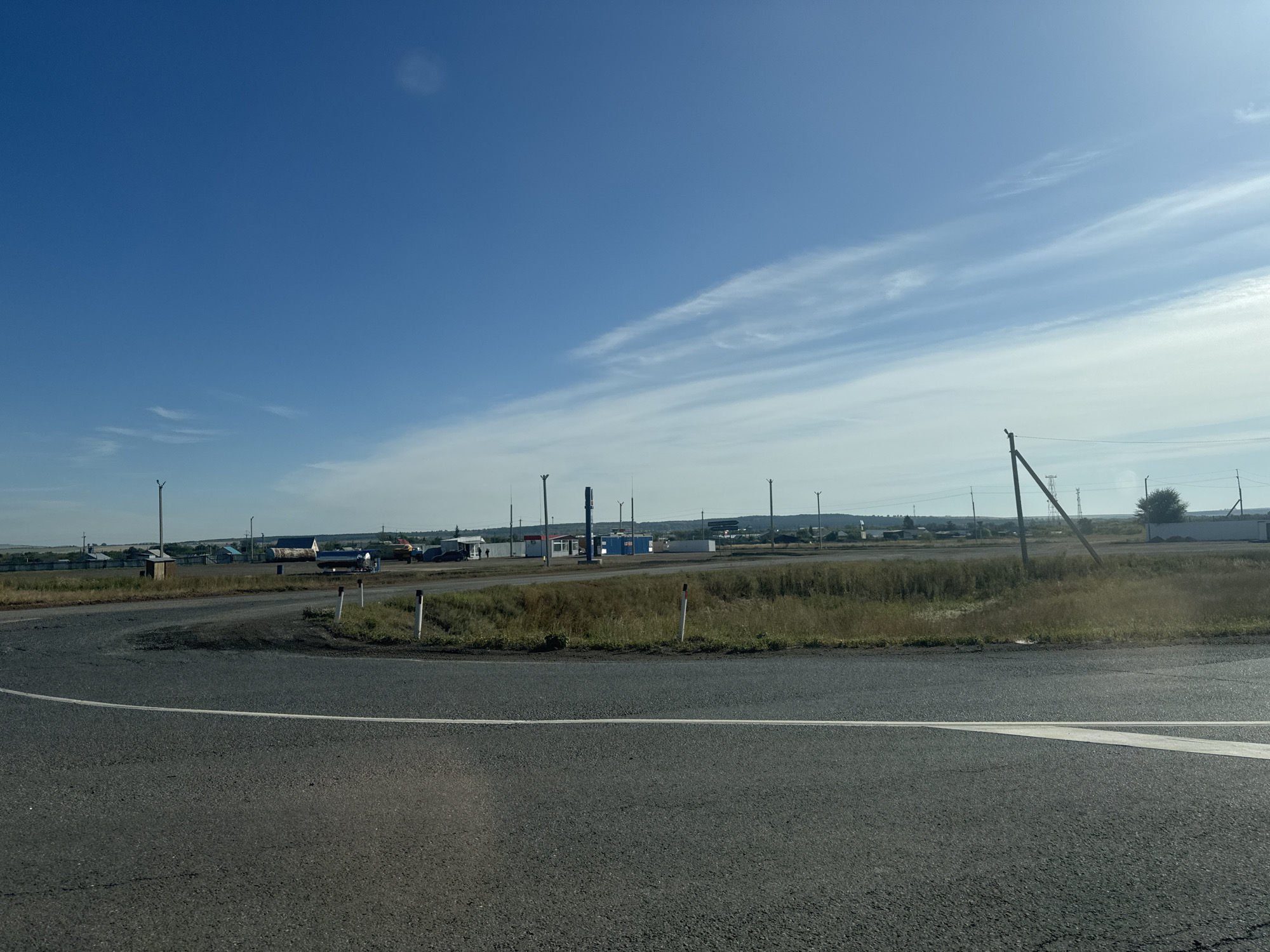 The image shows a rural road intersection with a small settlement visible in the background. The sky is clear blue with a few wispy clouds. In the foreground, there is an asphalt road with a white line marking. On the right side of the road, there is a grassy embankment with a few white posts. In the middle ground, there are several buildings that appear to be part of a gas station or rest stop. There are also some vehicles and utility poles. The settlement in the background consists of houses and fences, with rolling hills in the distance. The overall impression is a quiet, remote location on a sunny day.