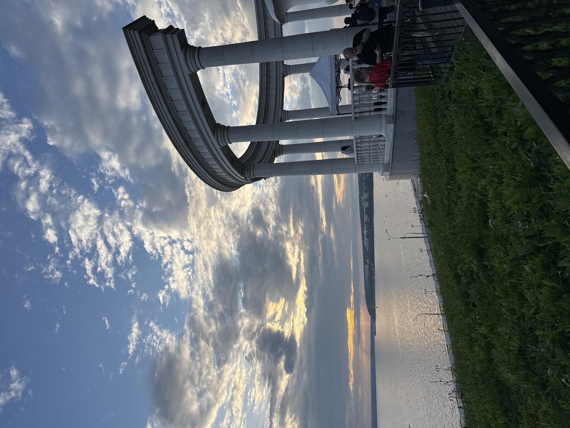 The image depicts a scenic view of the Izhevsk Pond in the city of Izhevsk, Russia, during what appears to be a sunset. In the foreground, there is a lush green grassy slope leading down towards the water. A white, open-air rotunda with classical columns and an ornate railing is visible on the right side of the image, overlooking the pond. Several people are standing on the promenade area within the rotunda, some interacting with each other. One person in the foreground, a child, is wearing a red shirt and looking towards the left. A man next to the child is looking down at his phone. A white canopy tent is set up behind the railing. The Izhevsk Pond stretches out to the horizon, reflecting the warm hues of the setting sun. The sky is a dramatic mix of blue and gray clouds, with sunlight breaking through in some areas, creating a radiant effect. The opposite shore of the pond is lined with trees and buildings, indicating the presence of the city. The overall atmosphere is tranquil and picturesque, capturing the beauty of the waterfront at dusk.