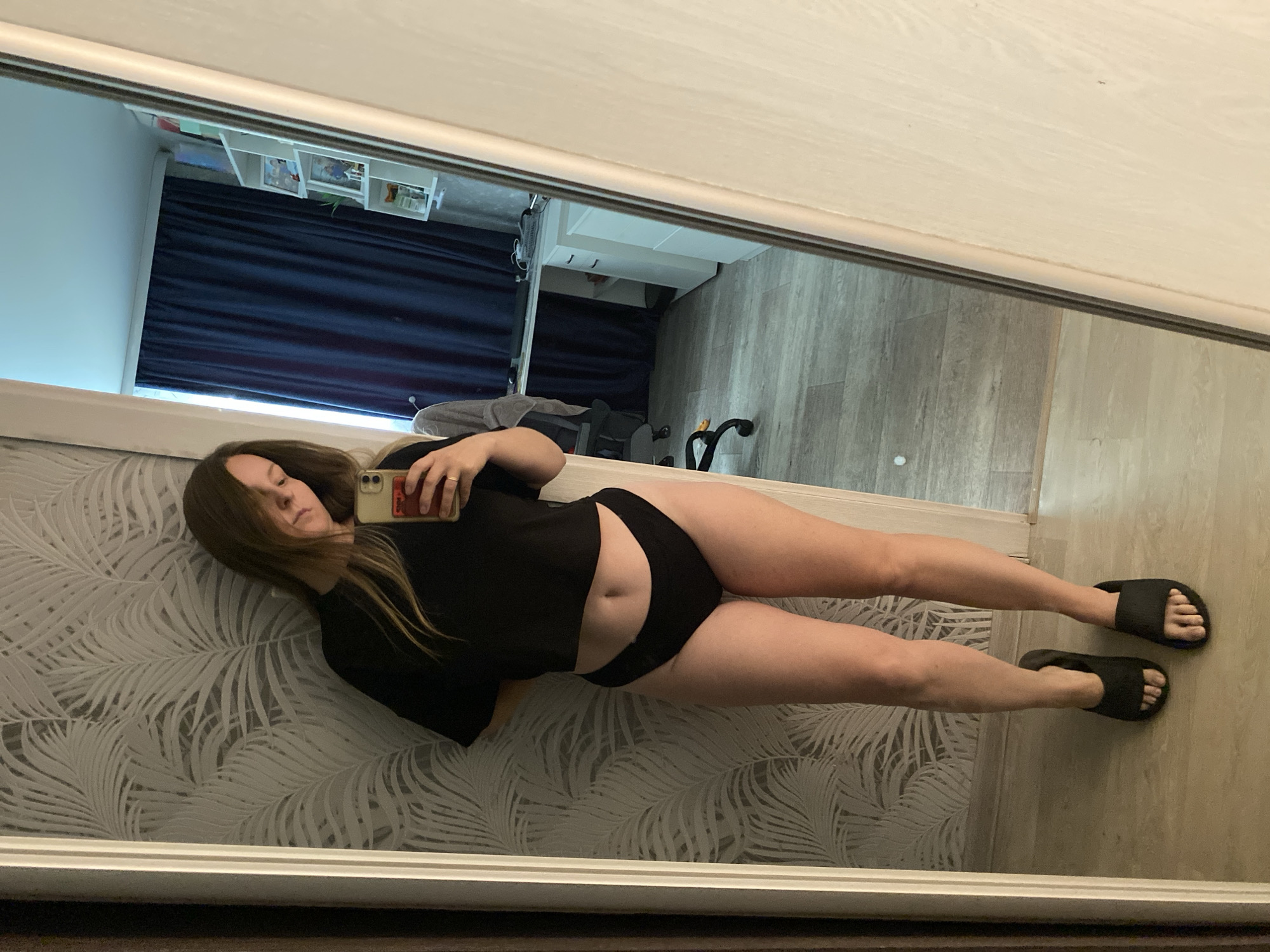 A woman is lying on a patterned bed while taking a selfie in a mirror. The mirror reflects a view of a room with dark blue curtains, a white shelf, and grey wood-look flooring. The woman is wearing a black t-shirt and black underwear and black slides. The location is a room interior; information about the city of Noviy, Russia, is not apparent from the image.