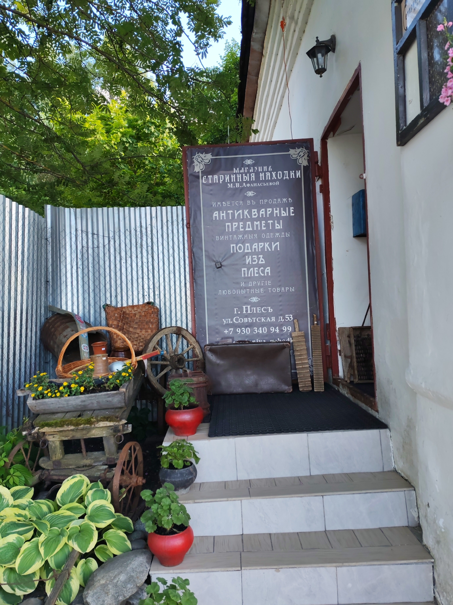 This media file depicts the exterior of a shop in Plyos, Russia. The scene is set outdoors, likely during the daytime with bright sunlight filtering through trees. The building appears to be old, with a white facade and a dark-trimmed doorway.
The shop's entrance is adorned with a large, dark sign that displays text in Russian. The sign advertises "MAGAZINE 'OLD FINDS' OF M.B. AFANASYEVA", and further details that they sell "ANTIQUARIAN ITEMS", "VINTAGE CLOTHING", "GIFTS FROM PLYO'S", and "OTHER CURIOUS GOODS". The address is listed as "G. PLYO'S, ul. Sovetskaya d. 53", and a phone number "+7 930 340 94 99" is also visible.
In front of the shop, there are several rustic objects arranged to create an inviting atmosphere. A wooden cart with a planter box filled with yellow flowers sits near a corrugated metal fence. A large wooden barrel, woven baskets, and an old wooden wheel are also present, adding to the antique aesthetic. Potted plants, including geraniums in red pots, are placed on the steps leading up to the entrance. A vintage leather suitcase is laid out on the ground in front of the steps. To the right of the entrance, a blue mailbox is mounted on the wall.
The overall impression is one of a charming, eclectic antique shop with a welcoming and nostalgic ambiance. The presence of greenery and flowers suggests a pleasant, possibly warm, day.