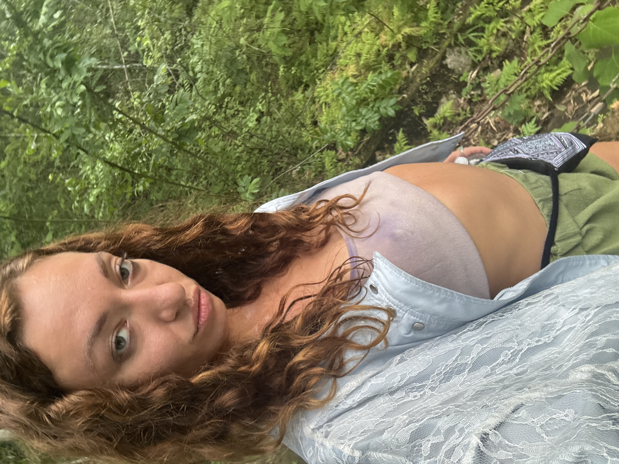 A woman with long, curly brown hair and a pale complexion poses for a selfie outdoors. She is wearing a light blue lace jacket over a light purple bralette and olive green shorts. She has a serious expression and is looking directly at the camera. The background is a lush, green forest, suggesting a natural, possibly remote, setting. The image does not provide specific details about the city of Xóm Khán, Vietnam, or any activities taking place.