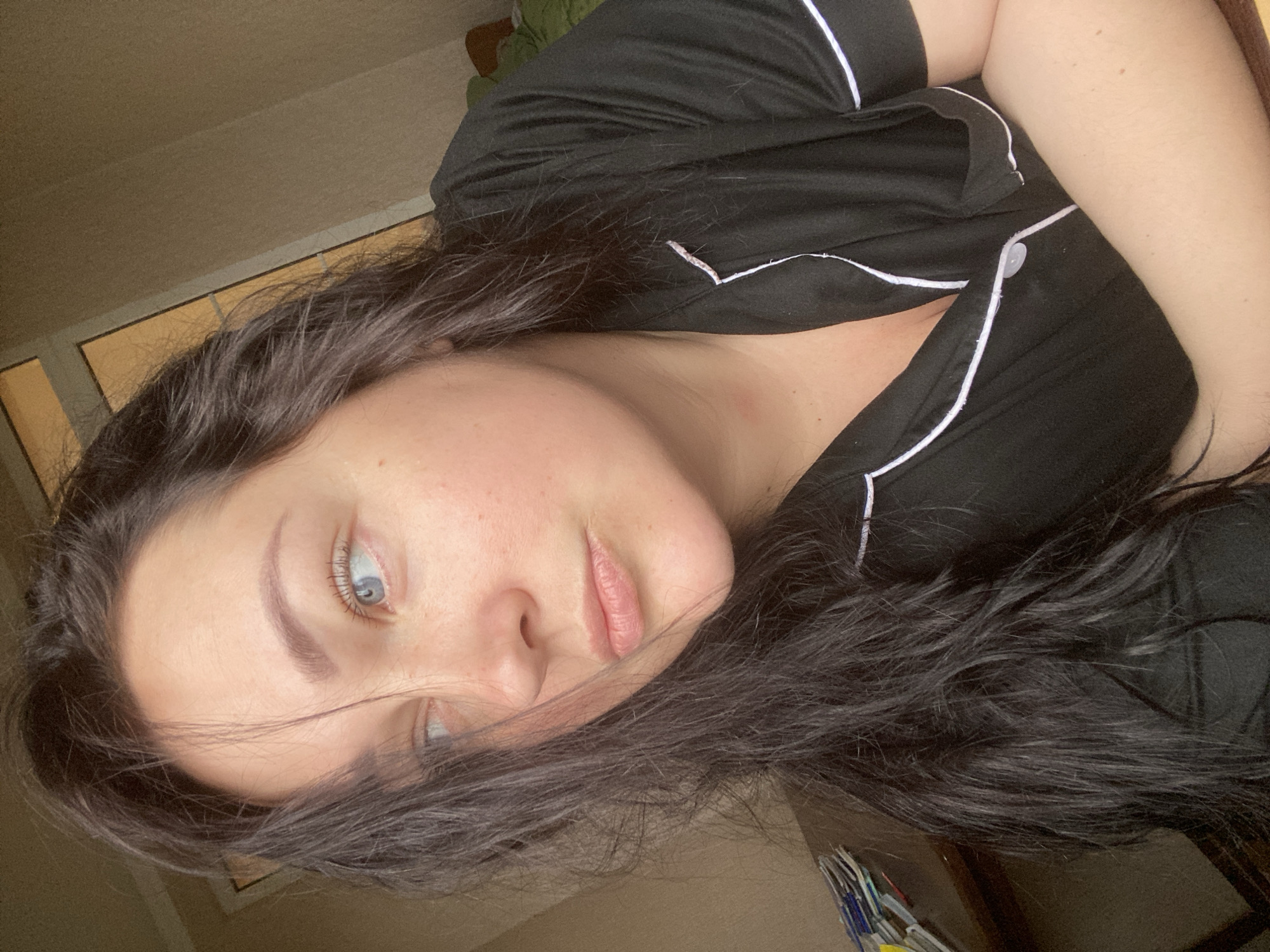A woman with long, dark hair is seen wearing black pajamas with white piping. She has fair skin and blue eyes. The background appears to be an interior space, possibly a bedroom, with a window visible. Location information is not discernible from the image.