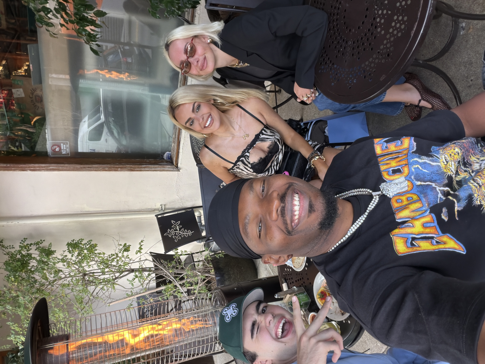 The image shows a group of four people taking a selfie at an outdoor dining area. Two blonde women are seated at a dark metal table. One is wearing a black blazer and the other a zebra-print top. Two men are taking the selfie. One is wearing a black shirt with a graphic design and a black durag, and the other is wearing a green baseball cap. A metal outdoor heater is visible in the bottom left corner. Dishes with food are visible on the table. Through a nearby window a commercial vehicle is visible. The location is likely in West Hollywood, United States of America.