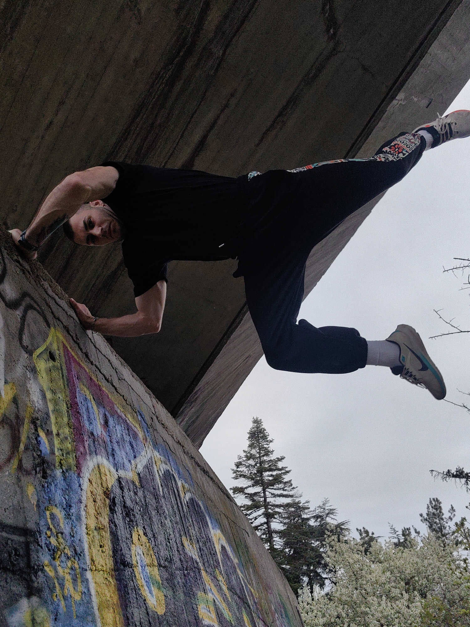 A man is suspended from a concrete structure, likely a bridge or overpass, in Varna, Bulgaria. He is holding onto the edge of a graffiti-covered concrete wall with his hands. He is wearing a black t-shirt, dark pants with colorful patterns on the upper portion of his left leg, white socks, and light-colored sneakers. His body is parallel to the ground, and he is looking down at the viewer. The background features overcast skies and trees.