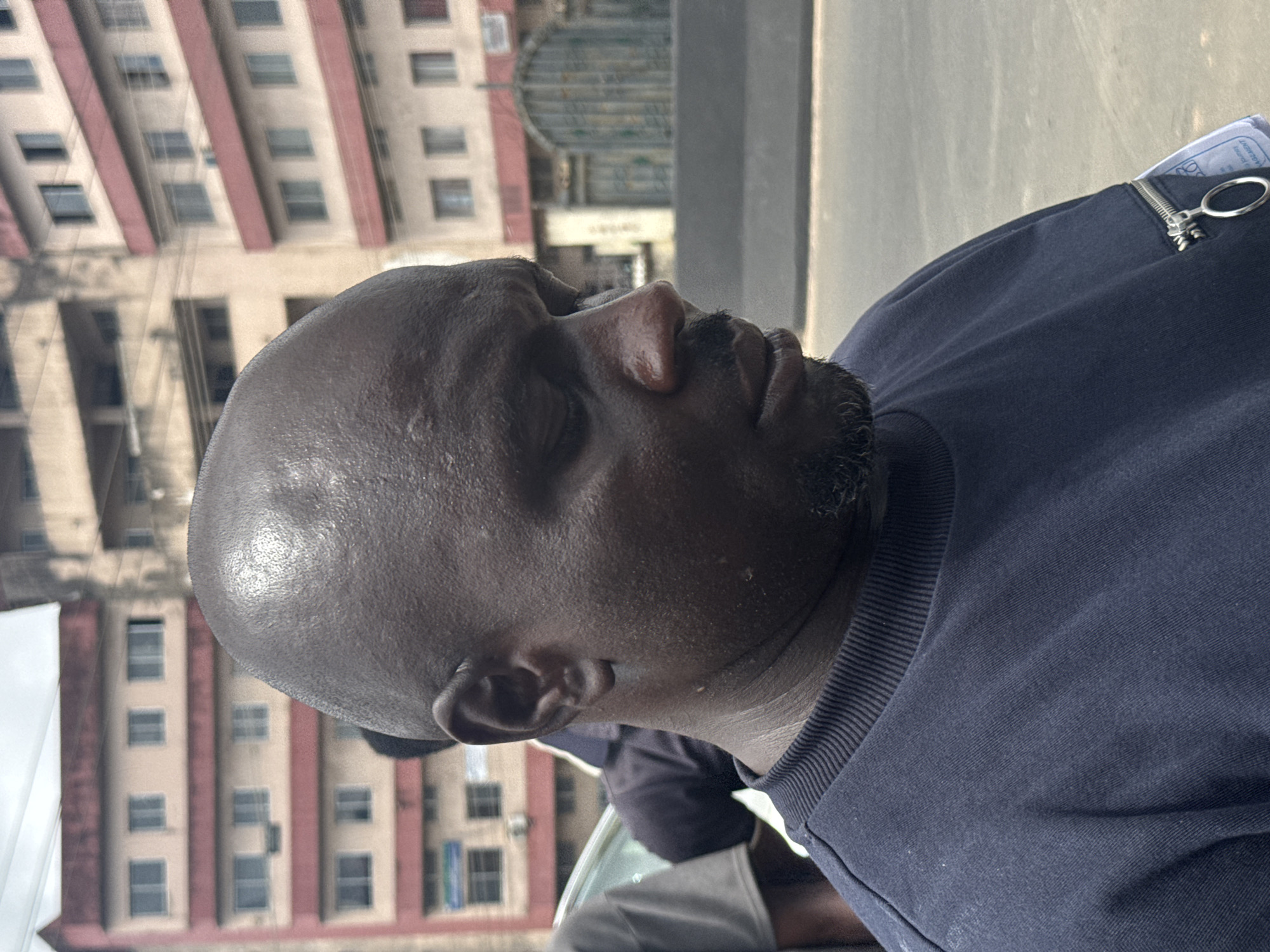 A close-up, medium shot captures a dark-skinned man with a shaved head, facing right with his eyes downcast, against a blurred urban background. He appears to be in his late 30s or early 40s, with weathered skin and a small goatee. He is wearing a navy blue sweatshirt with a ribbed collar. A silver zipper with a circular pull tab is visible on the left side of his shirt, along with a white tag. The background suggests an outdoor urban environment, possibly in Owerri, Nigeria, with multi-story buildings displaying beige and red facades. A grey barrier and a road are also visible. The lighting is diffused, indicating an overcast day.