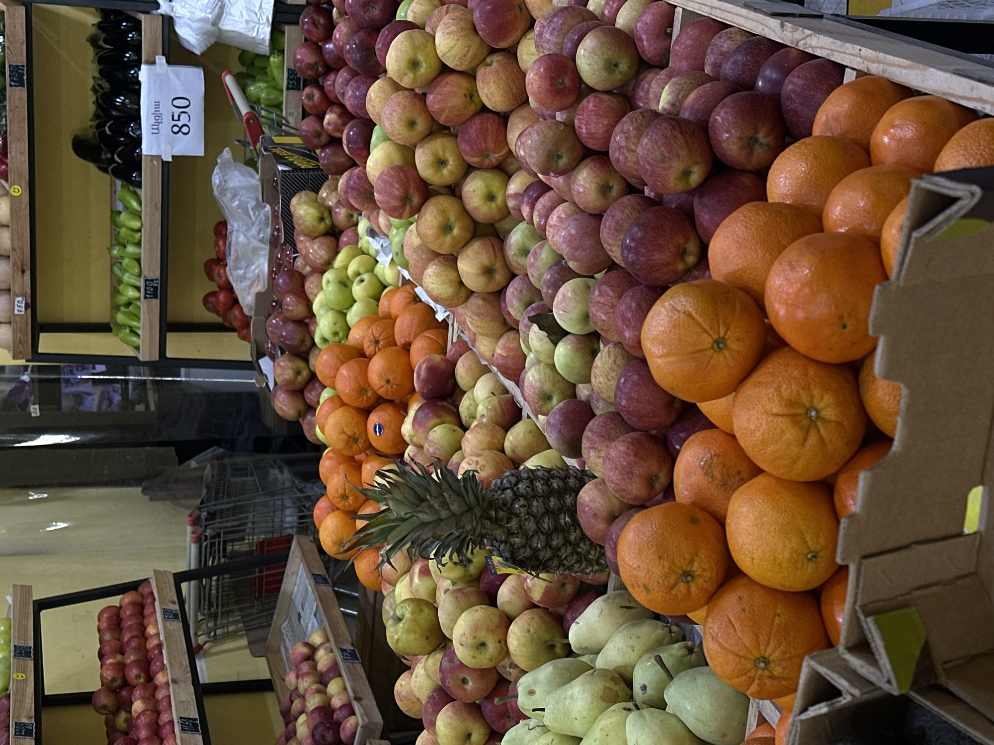 The image shows a grocery display in Yerevan, Armenia. The display features a variety of fruits and vegetables, including apples, oranges, pears, peppers, and eggplants. A pineapple is also visible. A price tag indicating "850" in Armenian script is attached near the peppers and eggplants. Shopping carts are visible in the background. The fruits and vegetables are arranged in stacked rows.