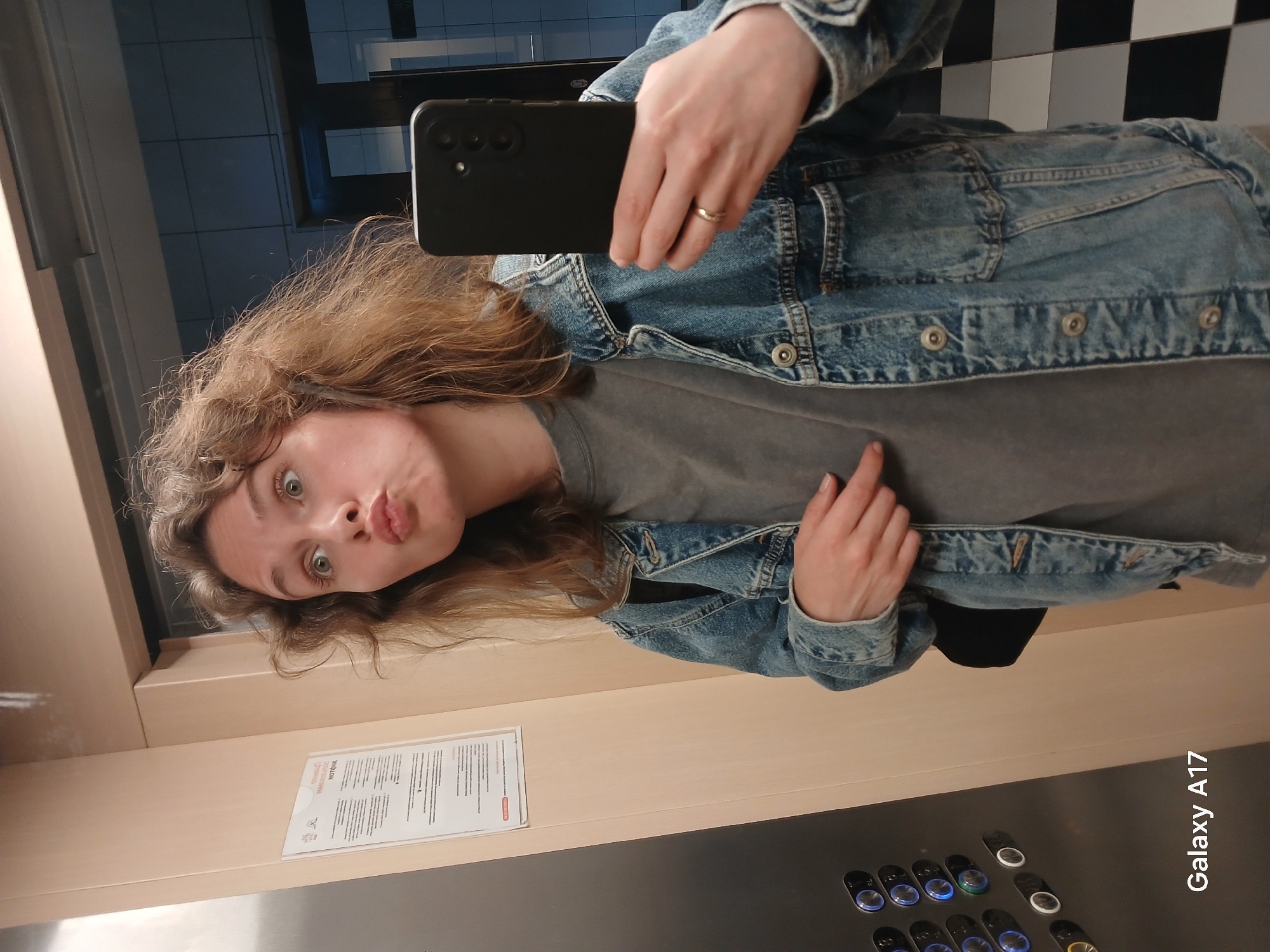 A female individual is depicted in a mirror selfie within an elevator in Yekaterinburg, Russia. She possesses wavy, medium-brown hair and light complexion, posing with wide eyes and pursed lips. She wears a grey t-shirt, a blue denim jacket, and a plain gold-colored ring on her right ring finger. Her right hand holds a black smartphone featuring a triple vertical camera setup, pointed towards the mirror. Her left hand is positioned with the index finger pointing towards her chest. The elevator interior consists of beige wall panels and a stainless steel control panel on the left, equipped with multiple illuminated blue circular buttons. A white document in a transparent holder, displaying Cyrillic text with the word "МОТОЧКИ" visible, is attached to the beige wall. The mirror's reflection reveals a tiled wall composed of dark grey/blue square tiles and a black-and-white checkered floor. The image metadata indicates it was captured by a "Galaxy A17" device.