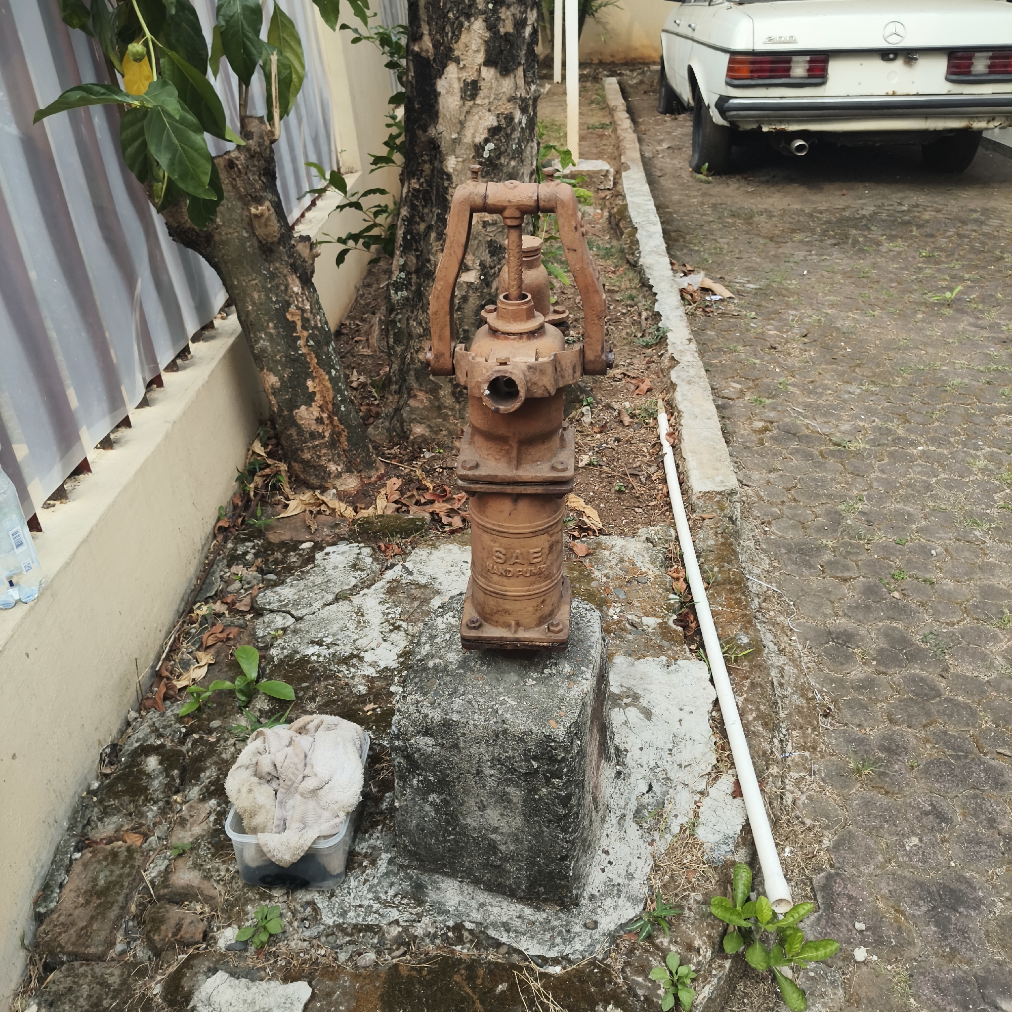 An abandoned, rusted manual water pump, identified by the inscription "SAE HAND PUMP," stands on a concrete pedestal in the foreground. It is surrounded by dry leaves, scattered dirt, and patchy vegetation. To the left of the pump, a clear plastic container holds a dirty, beige towel. A tree trunk with dark, rough bark is positioned behind the pump. Beyond the tree, a white, slightly weathered car is parked on a patterned stone pavement. A white PVC pipe runs along the edge of the pavement and the dirt area. In the background, a light-colored wall is visible, with a corrugated metal fence or siding. The overall scene suggests an outdoor, somewhat neglected area, possibly a residential yard or alleyway in South Jakarta, Indonesia. There are no people present in the image.
