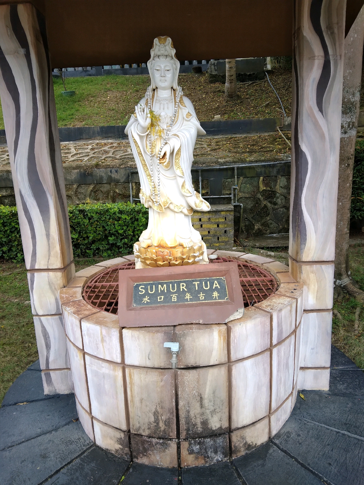 The image shows a statue of a woman in a white and gold robe, possibly a deity, standing on a lotus pedestal within a stone well structure. The well has four decorative pillars, painted with a wavy, abstract pattern of black, white, and beige. A dark brown plaque with gold lettering is mounted on the front of the well, reading "SUMUR TUA" in Roman script, and what appears to be Chinese characters below it. A spigot is visible on the side of the well. The well is surrounded by a grassy area and some low green bushes. In the background, there is a stone wall and more greenery. This scene is located at the Vihara Duta Maitreya temple in Batam, Indonesia, specifically in the area of the wishing well.