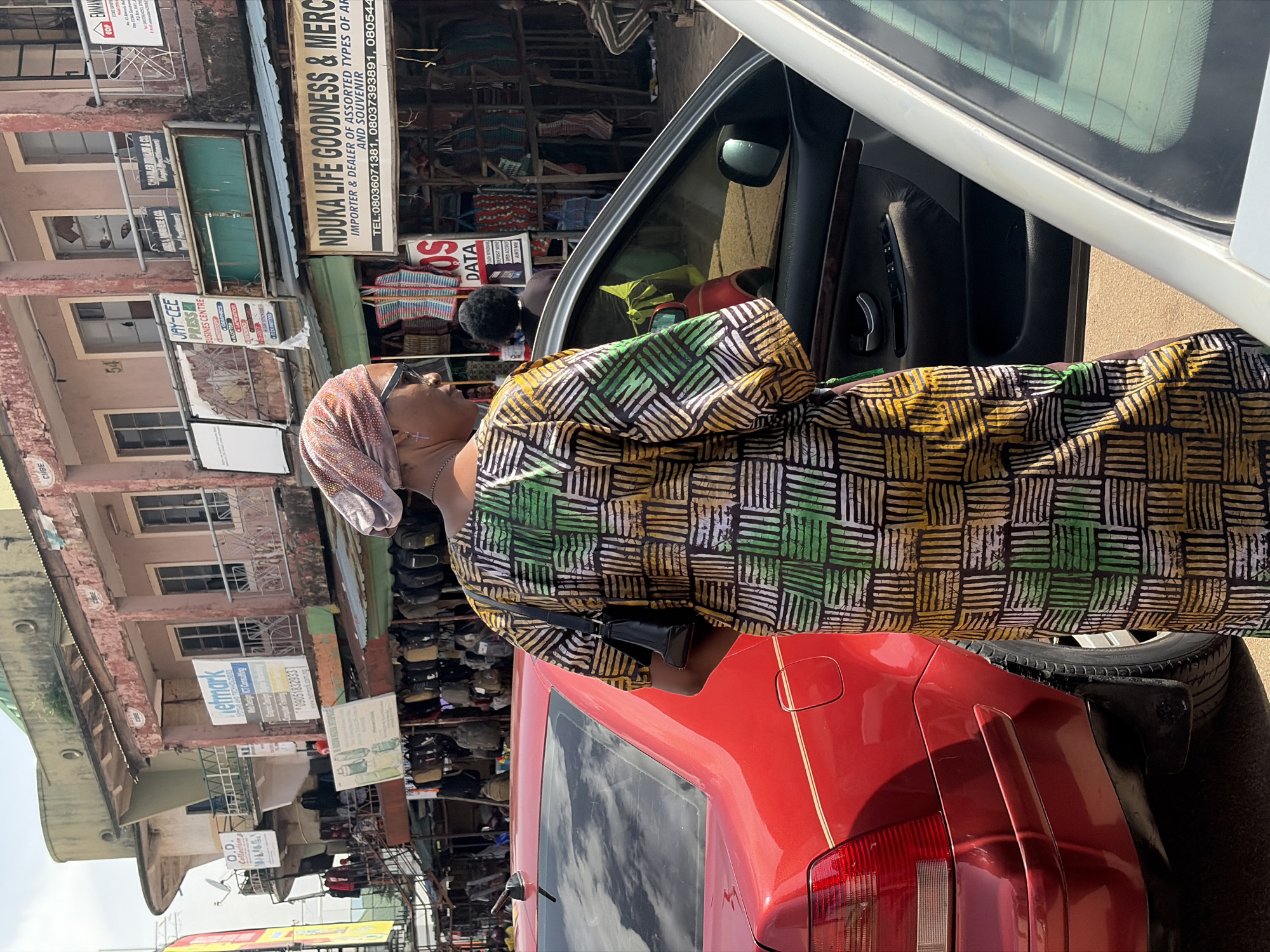 The image depicts a bustling street scene in Jos, Nigeria. A woman wearing a vibrantly patterned traditional outfit and a headscarf is seen from the side, leaning towards the open door of a red car. She appears to be either entering or exiting the vehicle. The background reveals a row of shops with various signs, suggesting a commercial area. The weather appears to be bright and sunny, indicating daytime.
Visible text includes:
- "NDUKA LIFE GOODNESS & MER..." on a sign above a shop.
- "JAY-CEE PRESS BUSINESS CENTRE" on a sign.
- "Netmark" on a blue sign.
- "O.D. Collections M&K, S.E." on a sign.
- "COS DATA" on a red sign.
- A sign with "WEB DESIGN, IT CONSULTING".
- A phone number "08036071381, 08037393891, 080544..."
The scene is lively and indicative of everyday commerce and activity on a Nigerian street. The woman's attire suggests a cultural context, and the presence of multiple shops implies a marketplace or commercial district. The overall impression is one of vibrant daily life.