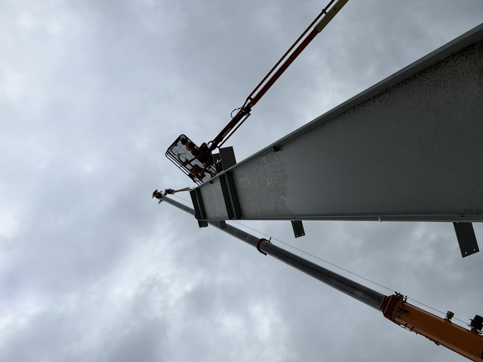 In Janowice, Poland, a construction project is underway, focusing on the installation of a large, grey steel beam. The beam is oriented vertically and angled upwards, dominating the foreground. A crane, with its bright orange boom extending from the bottom left of the frame, is actively involved in the operation, its cable attached to the beam. Further up, a cherry picker is positioned against the steel structure, its basket at the top, with two individuals visible inside, seemingly working on the beam. The sky above is overcast with heavy clouds, suggesting potentially unfavorable weather conditions for construction. The overall scene depicts a critical phase of building assembly, with heavy machinery and personnel working at height.