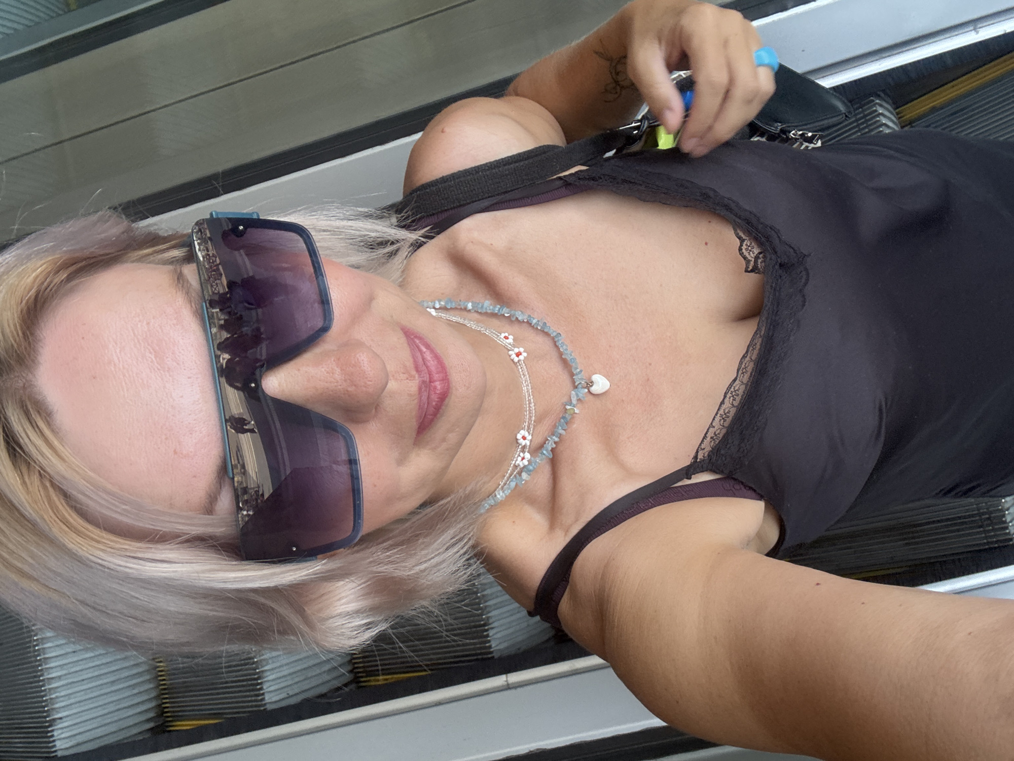 A woman with blonde hair and sunglasses takes a selfie on an escalator. She is wearing a black camisole with lace trim and a necklace. The escalator appears to be in a public place, possibly a shopping mall or transportation hub. The background is blurred, but suggests an indoor environment. The reflection in her sunglasses shows a group of people walking. There are no other discernible details about the location or specific actions. The overall impression is of a casual snapshot taken in a metropolitan setting.