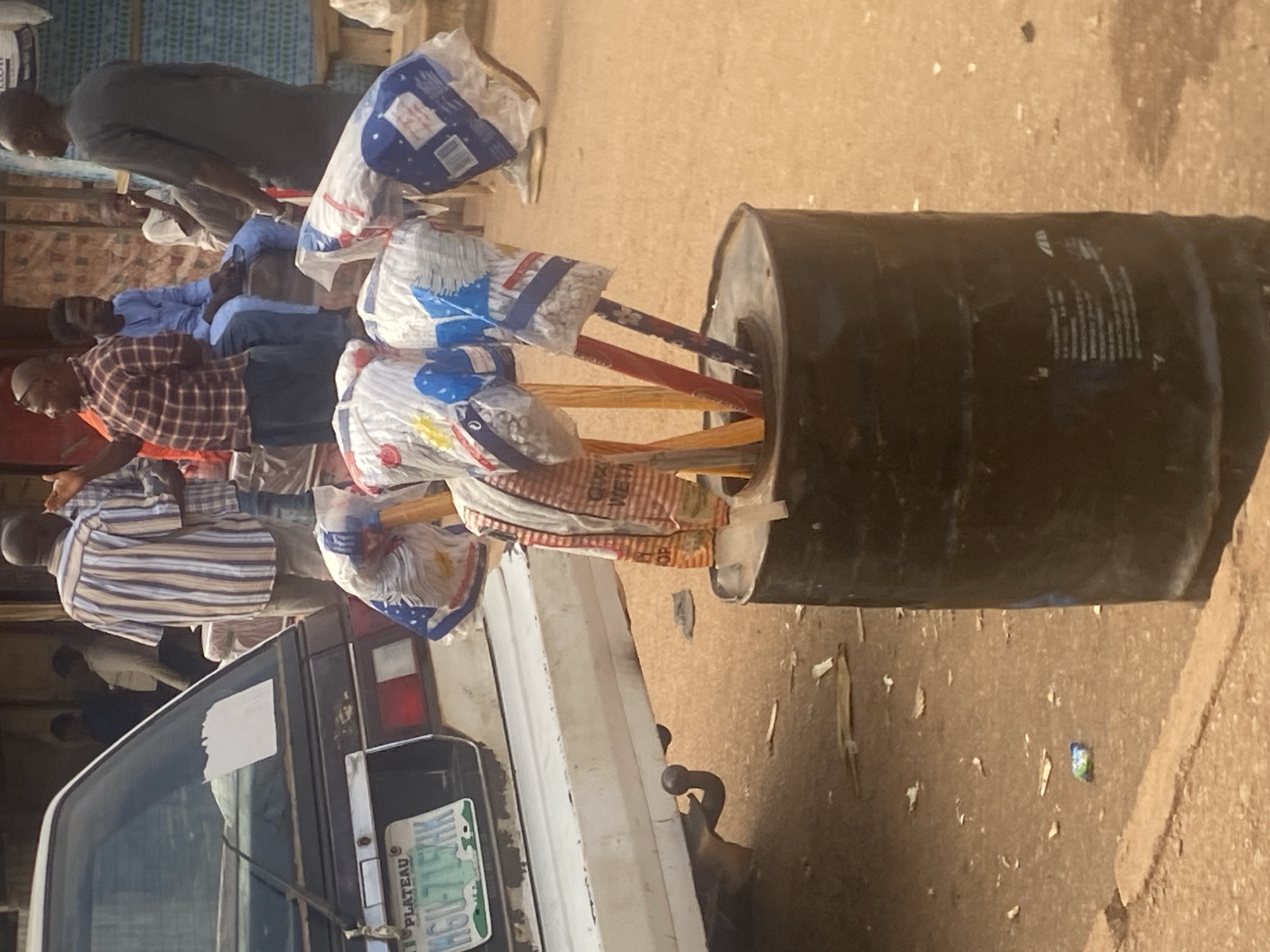 In Bukuru, Nigeria, a scene unfolds near a parked car, possibly a station wagon with a license plate reading "PLATEAU HGU-715HK". Several individuals are present, including a man in a blue outfit, another in a plaid shirt, and a man in a striped shirt. In the foreground, a black barrel serves as a makeshift stand for a collection of items, each wrapped in plastic. These items appear to be fireworks, with colorful sticks protruding from the barrel and plastic packaging featuring designs that suggest pyrotechnics, some with blue accents. The ground is dirt and debris. In the background, there are structures that appear to be market stalls or shops.