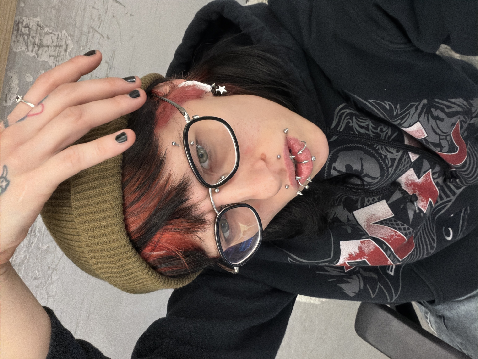 A person with black and red hair is wearing a green beanie, black-rimmed glasses, and a black hooded sweatshirt with a red and white design. They have multiple facial piercings on their lips and eyebrows, and a star earring in their ear. Their fingernails are painted black, and they have tattoos on their hand. The background appears to be a neutral grey wall. The location in Sofia, Bulgaria is not determinable from the image.