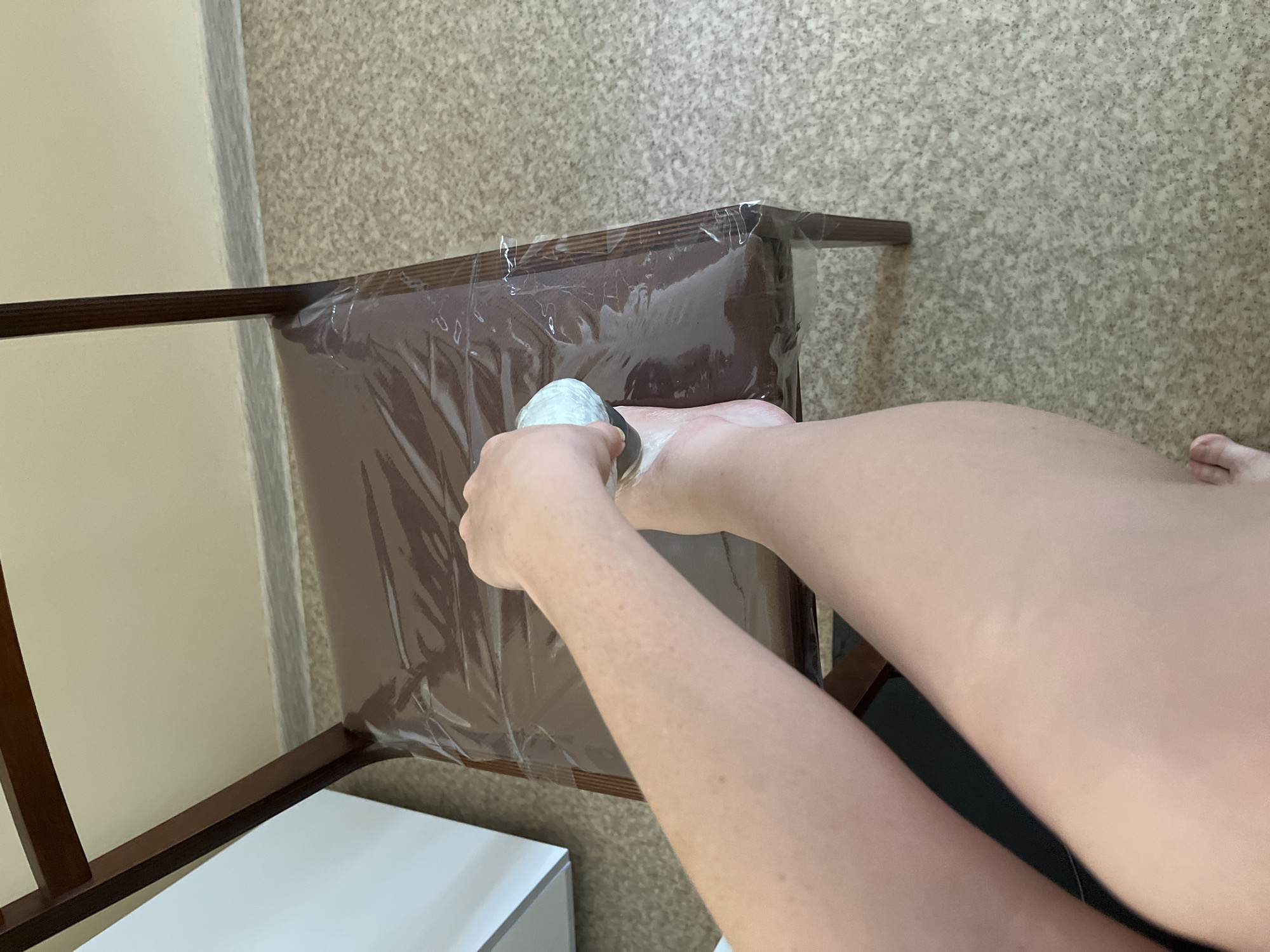 An overhead shot depicts a person, likely female, seated and using a gua sha tool on their foot, which rests on a chair covered in clear plastic. The chair is brown wood with a white section visible beneath the seat. The background consists of a light beige wall on the left and a beige speckled floor on the right. There is no visible indication of the city of Chaikovsky, Russia.