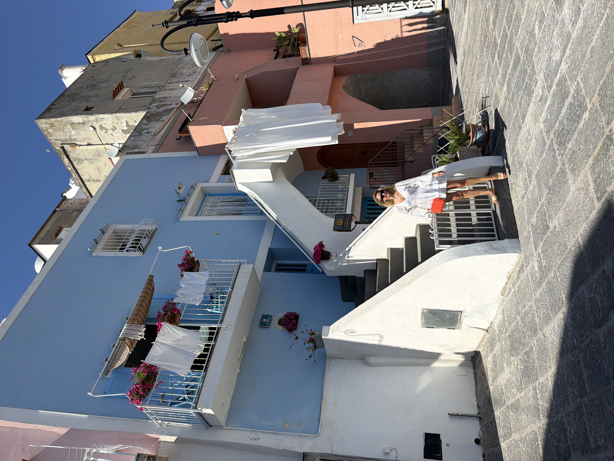 A woman in a white dress stands on outdoor steps near colorful buildings in Procida, Italy. The buildings are painted in shades of blue, pink, and yellow. Laundry hangs from the balconies of the blue building. A stone-paved street runs alongside the buildings.