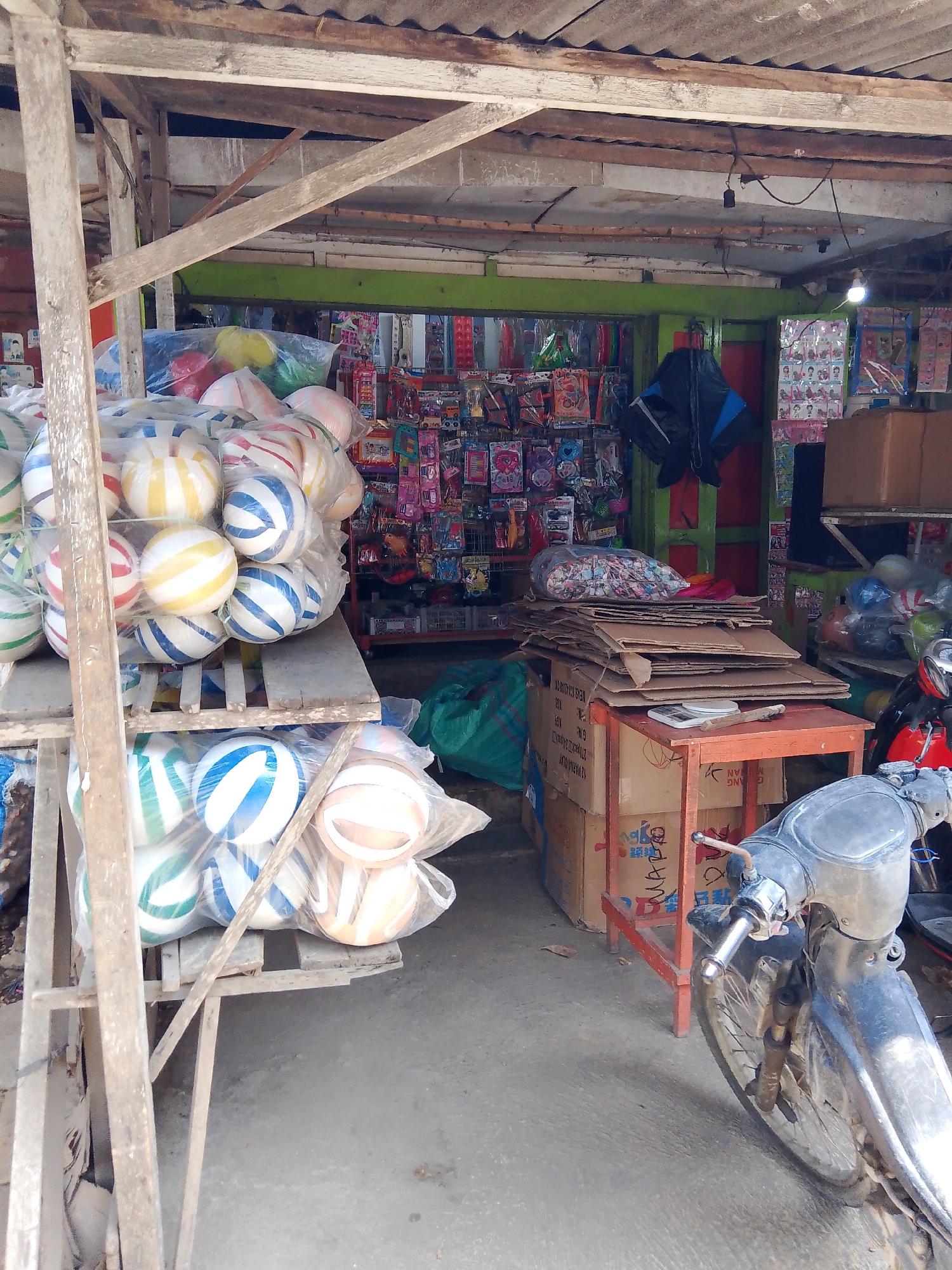 The image depicts an outdoor market stall in Gedangan, Indonesia, with a focus on a collection of inflatable balls. The stall has a rustic wooden frame with a corrugated metal roof. Various goods are displayed on shelves and in plastic bags, including a large assortment of colorful striped inflatable balls in the foreground and midground, some in plastic bags. Behind the balls, the stall's interior is visible, featuring shelves stocked with packaged items, possibly toys or novelty items, with colorful packaging. There are also several cardboard boxes stacked, and a green tarp is visible on the ground. A red metal table is present, with more stacked cardboard on top. A motorcycle, mostly silver, is parked in the foreground on the right side of the image. The ground is concrete. No people are visible in the image.