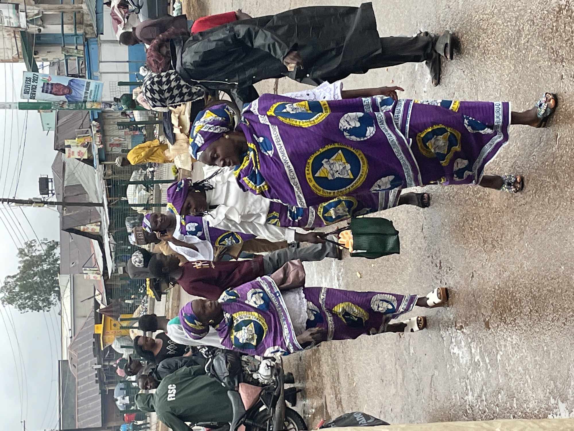 In Bukuru, Nigeria, a bustling street scene unfolds under an overcast sky, suggesting recent or ongoing rain as evidenced by the wet pavement reflecting light. A group of people are gathered, some walking, others standing. The central focus is on several women adorned in vibrant purple attire, intricately patterned with circular emblems featuring depictions of globes and possibly figures or symbols within them, along with text that includes "ECWA WOMEN-FELLOWS" and "BUKURU-D".
One woman in the foreground, wearing a purple headwrap and a matching patterned robe over white clothing, walks with purpose. She carries a brown handbag over her shoulder and wears distinctive platform sandals with white straps and wooden soles. To her left, another woman in similar purple attire also walks, her patterned robe partially obscuring her white clothing, and she wears flat sandals. A third woman, further left and slightly behind, is also dressed in the same distinctive purple pattern.
In the background, other individuals are visible. A person in a green hooded raincoat with "FISC" printed on the back stands near a motorcycle. To the right, a man in a black raincoat walks away from the camera, and another man in a maroon t-shirt is partially visible. Further in the background, market stalls with corrugated metal roofs line the street, and a political banner for "SELFLESS SERVICE 2027" featuring a man's portrait is attached to a pole. The environment appears to be a commercial area with buildings and infrastructure typical of a Nigerian town. The overall atmosphere is one of daily life and commerce, with a strong presence of cultural attire.