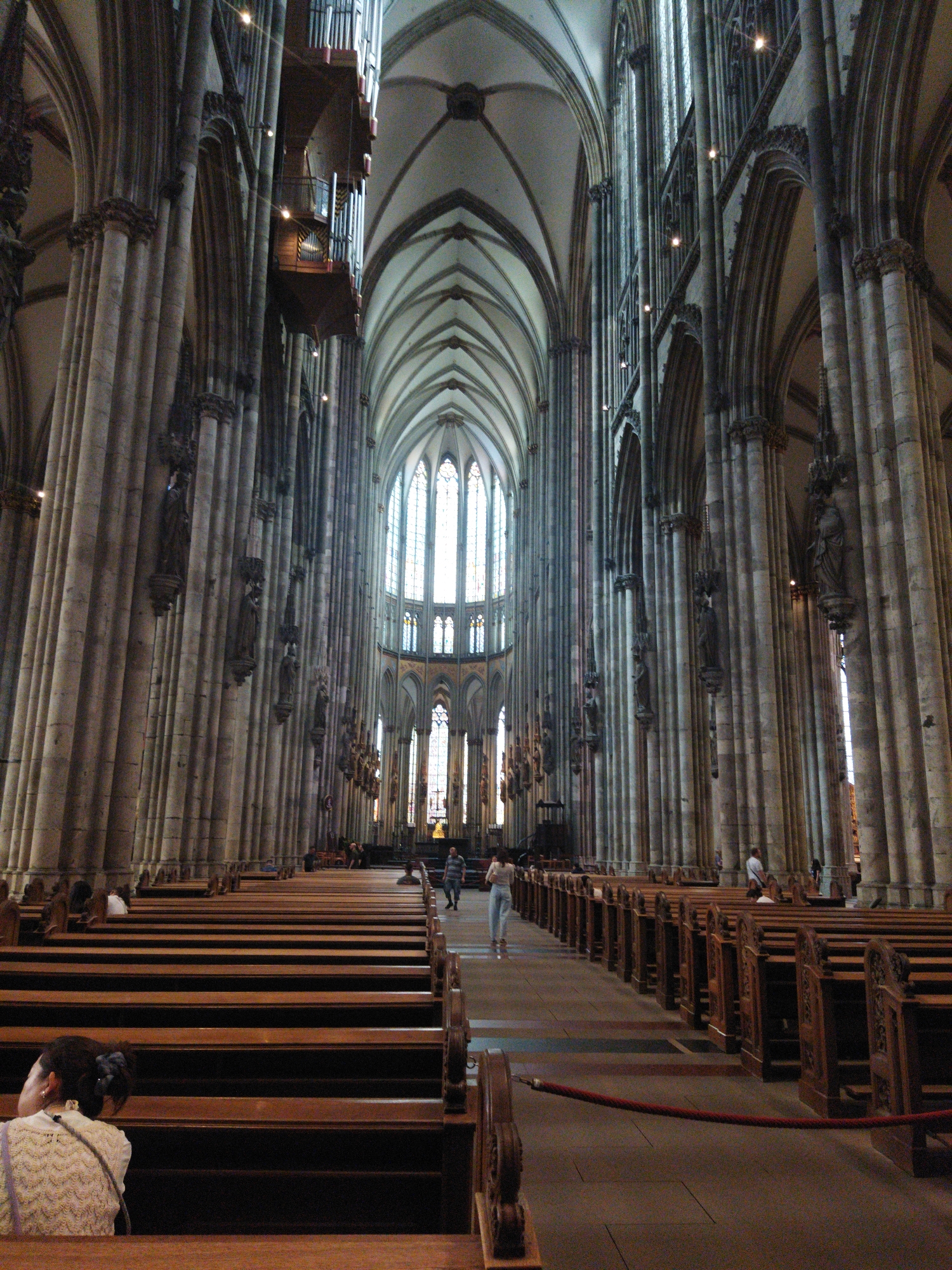 The image shows the interior of a grand gothic cathedral. Tall, ornate columns line the nave, which is illuminated by natural light filtering through large stained-glass windows. Rows of wooden pews stretch down the length of the cathedral. Several people are visible: a woman in the foreground facing away from the camera, another woman walking down the aisle, and a few individuals in the background. The architecture features high vaulted ceilings and intricate carvings. The organ is mounted high on the wall. The scene suggests a place of worship or a historical landmark. The image is taken inside the Cologne Cathedral, located in the city of Cologne, not Monheim am Rhein, Germany.