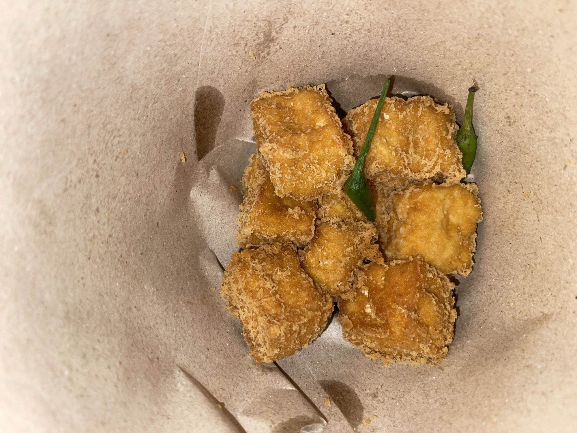 In Dewasari, Indonesia, a paper cone holds a pile of golden-brown, crispy fried tofu pieces. Two small green chilies are scattered amongst the tofu. The paper cone's texture is rough and brown, suggesting it's a common take-out container. The lighting in the image is warm and directional, casting shadows that highlight the texture of the food and the paper. The overall scene is a simple, close-up shot of a snack.