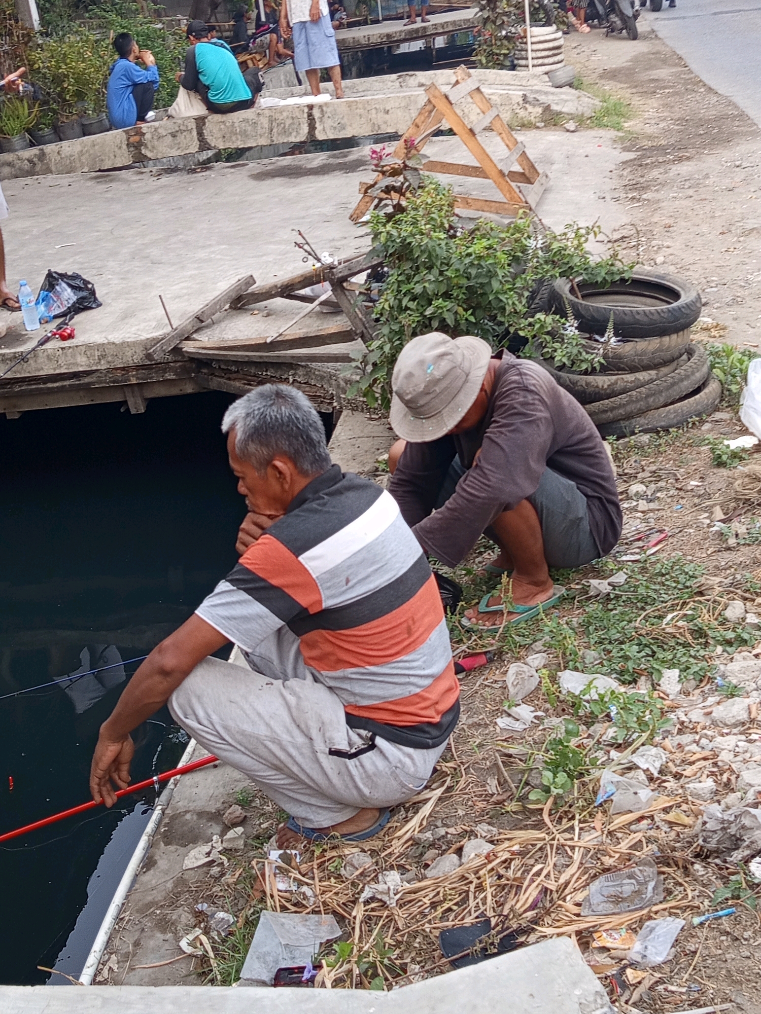 In Karawang, Indonesia, two men are seen fishing by a murky body of water. The man in the foreground, wearing a striped shirt and gray pants, is squatting and holding a red fishing rod. To his right, another man in a dark long-sleeved shirt and shorts, wearing a hat and flip-flops, is also squatting and appears to be tending to something on the ground. The environment around them is unkempt, with debris, dried vegetation, and stacked tires on the embankment. In the background, other individuals are visible, some sitting and others standing, on a concrete structure near potted plants. The scene suggests a casual fishing activity in a somewhat neglected urban area.