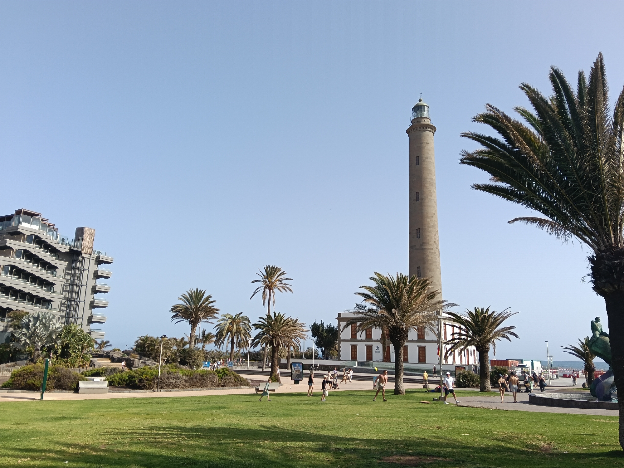 The image shows a sunny scene at Maspalomas, Spain, featuring the prominent Maspalomas Lighthouse. The lighthouse, a tall cylindrical structure with a green top, is centrally located with a white and red-striped building at its base. Lush green grass fills the foreground, interspersed with palm trees. Several people are seen walking, playing, and relaxing on the grass. A modern building with balconies is visible to the left, and a partially visible fountain sculpture with a seated figure is on the right. The sky is clear and blue. The scene suggests a popular recreational area near the beach.