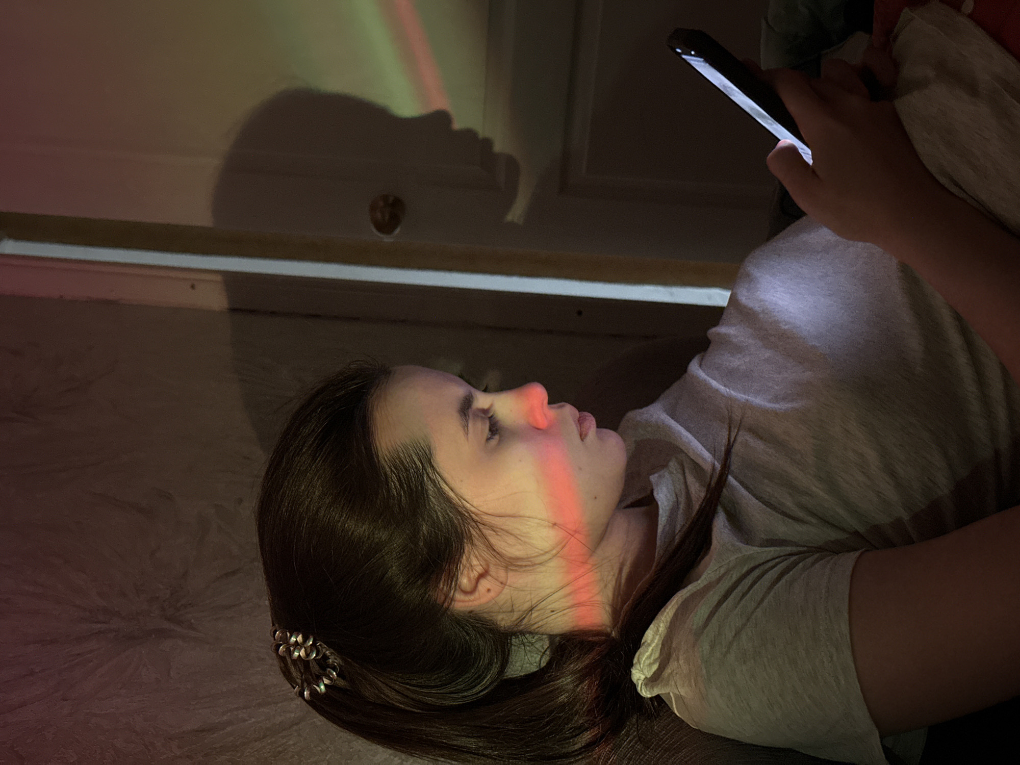 The image captures a young woman lying on her side indoors, engrossed in her smartphone. She is seen in profile, facing right, with her long dark hair spread around her. A decorative silver-toned hair clip is visible in her hair near her ear. She wears a light-colored, possibly grey, t-shirt.
The setting appears to be a dimly lit room, likely a bedroom or living area in Chaikovsky, Russia. Her right hand holds the smartphone, whose bright screen illuminates her hand, a portion of her face, and her clothing. A striking detail is a multi-colored light beam, primarily red, that cuts across her nose and cheek, extending onto the wall behind her. This beam also shows hints of green and yellow, suggesting it might be cast by a prism or a specialized light source.
On the wall in the background, a distinct shadow resembling a human profile or face is visible, alongside a circular object that could be a doorknob or part of a fixture. The low light and the woman's relaxed posture suggest the activity is taking place in the evening or at night. Her expression is calm and focused, indicating deep concentration on her phone's content. There is no visible text in the image.
